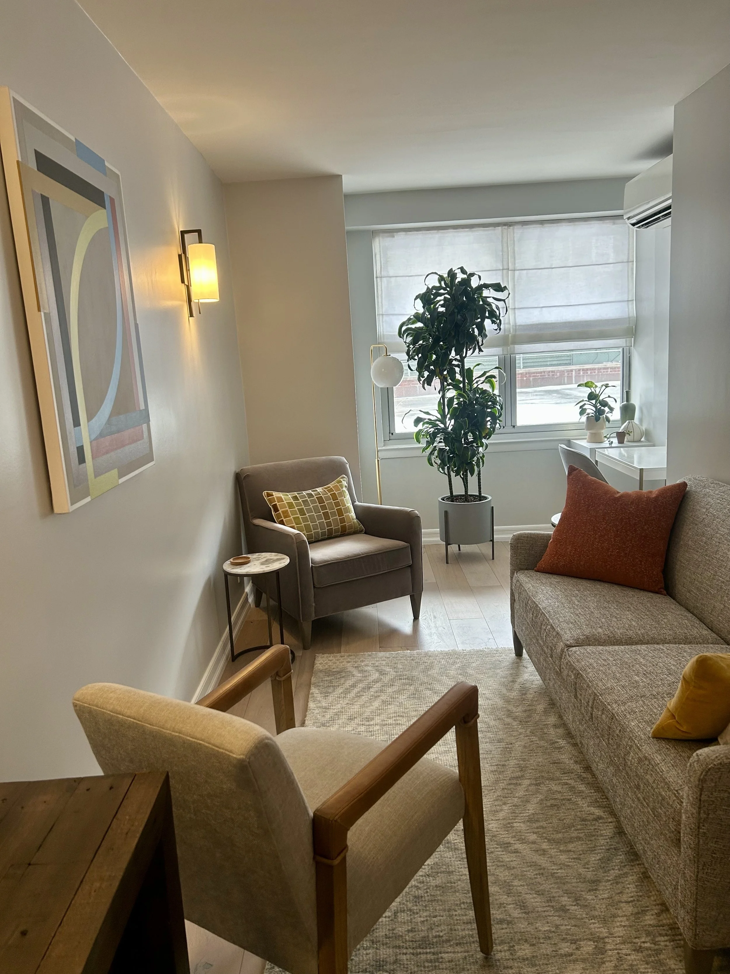 A view of Office Two in a therapy office for rent in Brooklyn, with a beige armchair, a gray sofa with orange and yellow pillows, a wooden side table, a tall potted plant,  windows with Roman Shades, and wall art with abstract geometric shapes.