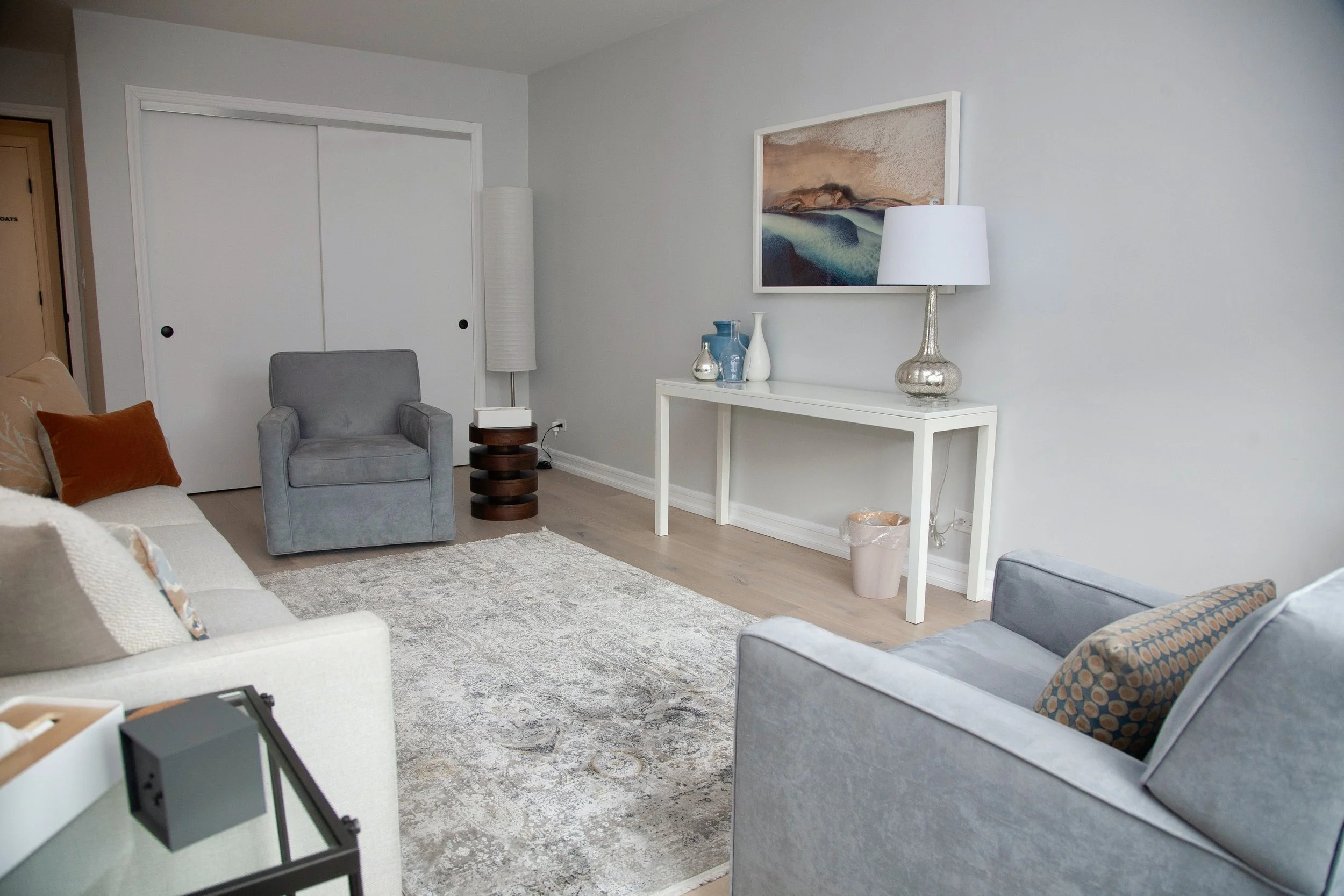Office Three in a therapy office for rent in Brooklyn, view of a beige sofa, gray armchair, patterned pillows,  rug, decorative vases on a white table, framed artwork on the wall, floor lamp, and closet with sliding doors.