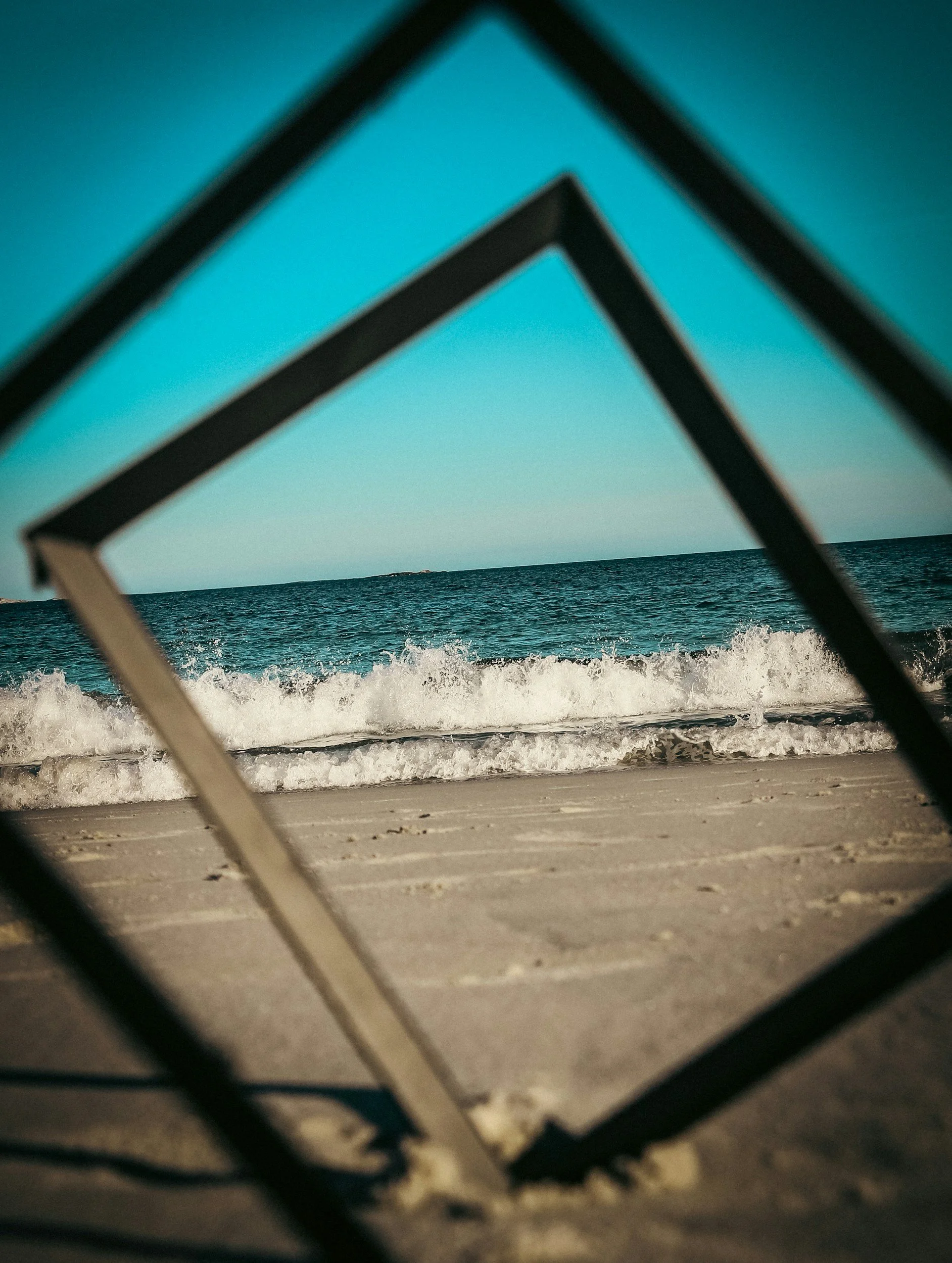 View of the beach through a geometric frame, showing sand, ocean waves, and a clear sky.