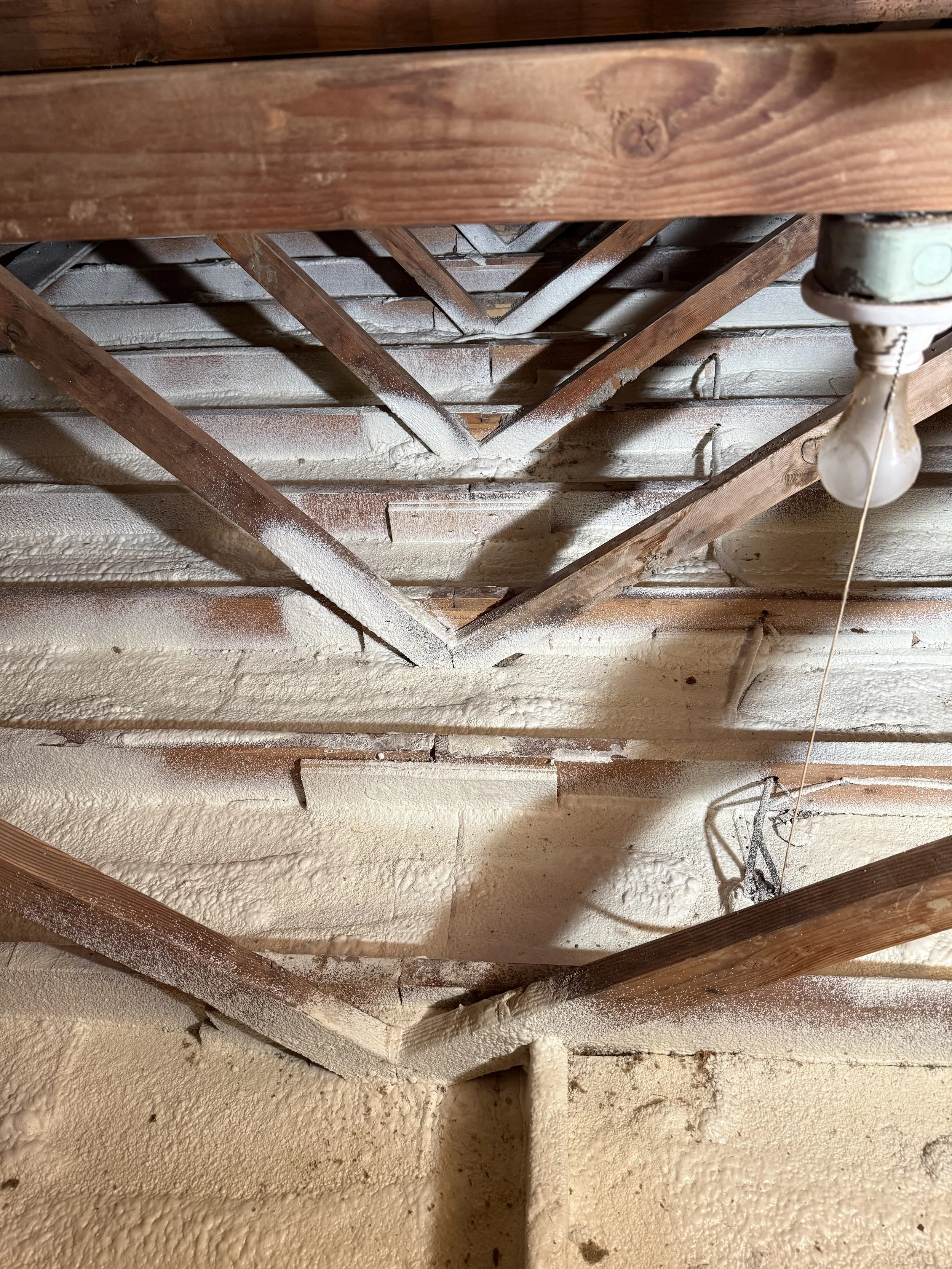View of a corrugated metal ceiling with wooden beams and a hanging light bulb, with some spray foam insulation on the ceiling.