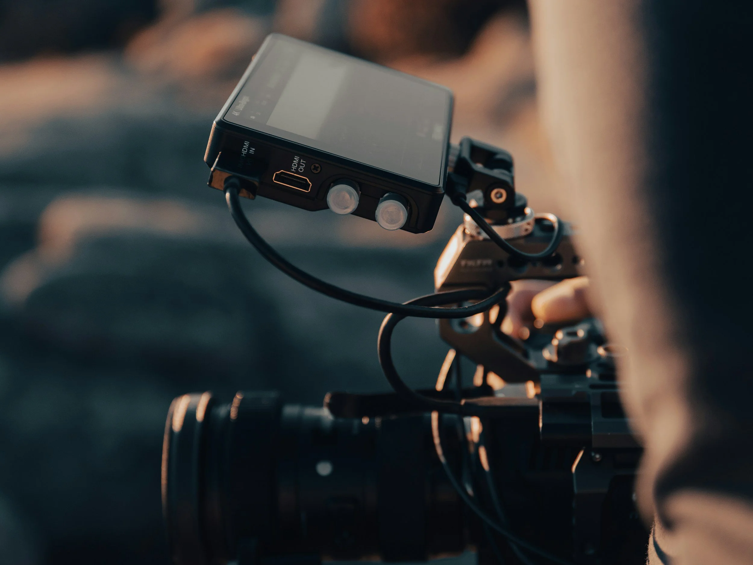 Close-up of a professional video camera with an attached monitor and cable, being operated outdoors during sunset.