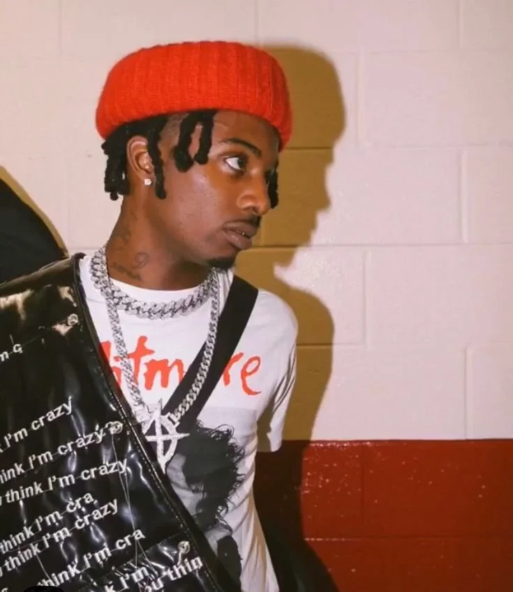 A young man with dreadlocks wearing a red beanie, multiple necklaces, earrings, a printed white t-shirt, and a black shiny jacket, standing against a white and red wall with his shadow cast on the wall.