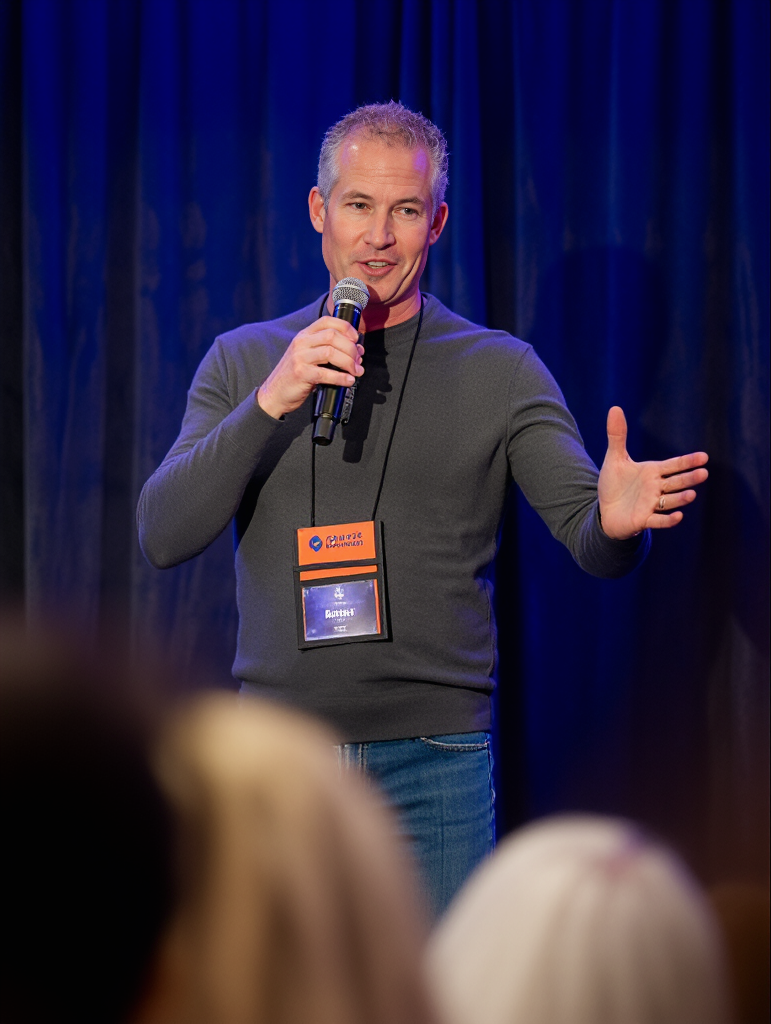 A man with short gray hair, wearing a gray long-sleeve shirt, speaking into a microphone on stage with a blue curtain background, gesturing with his right hand, and wearing a name badge.