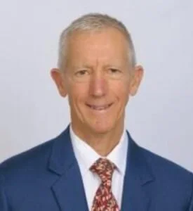 A smiling older man in a blue suit, white shirt, and patterned tie against a plain light background.