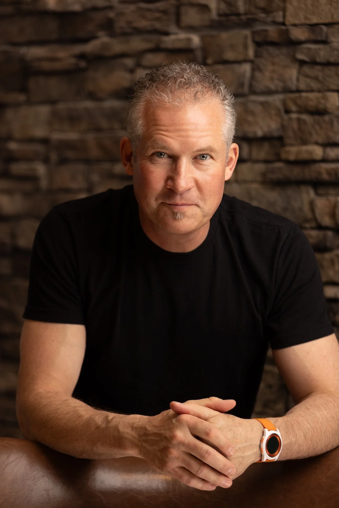 A man with curly gray hair and blue eyes, wearing a black t-shirt and an orange and white smartwatch, sits at a wooden table against a brick wall background.