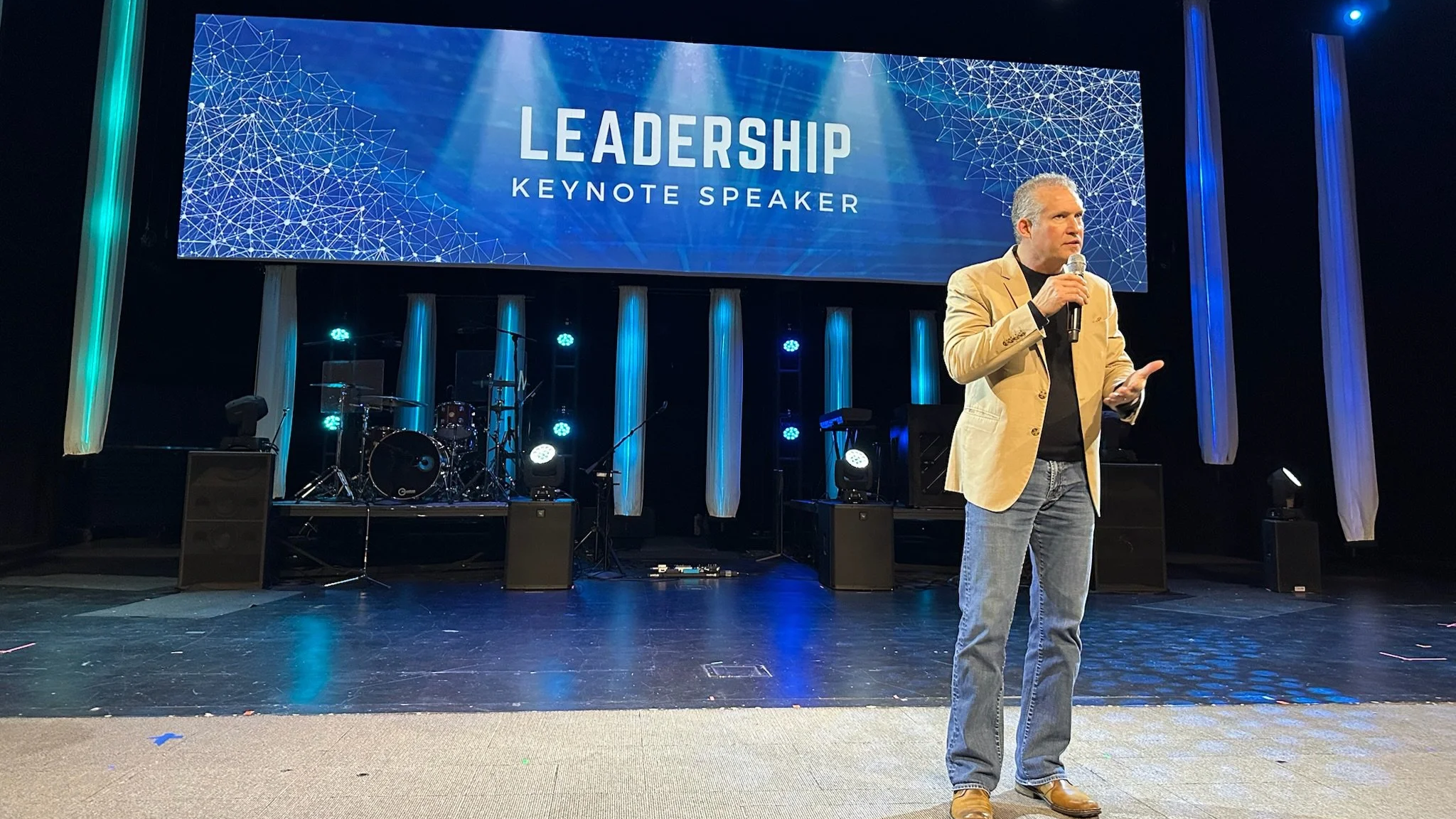 A man with gray hair wearing a tan blazer, black shirt, and jeans speaking into a microphone on a stage with a large screen behind him displaying the words "Leadership Keynote Speaker". The stage has musical instruments and blue lighting.