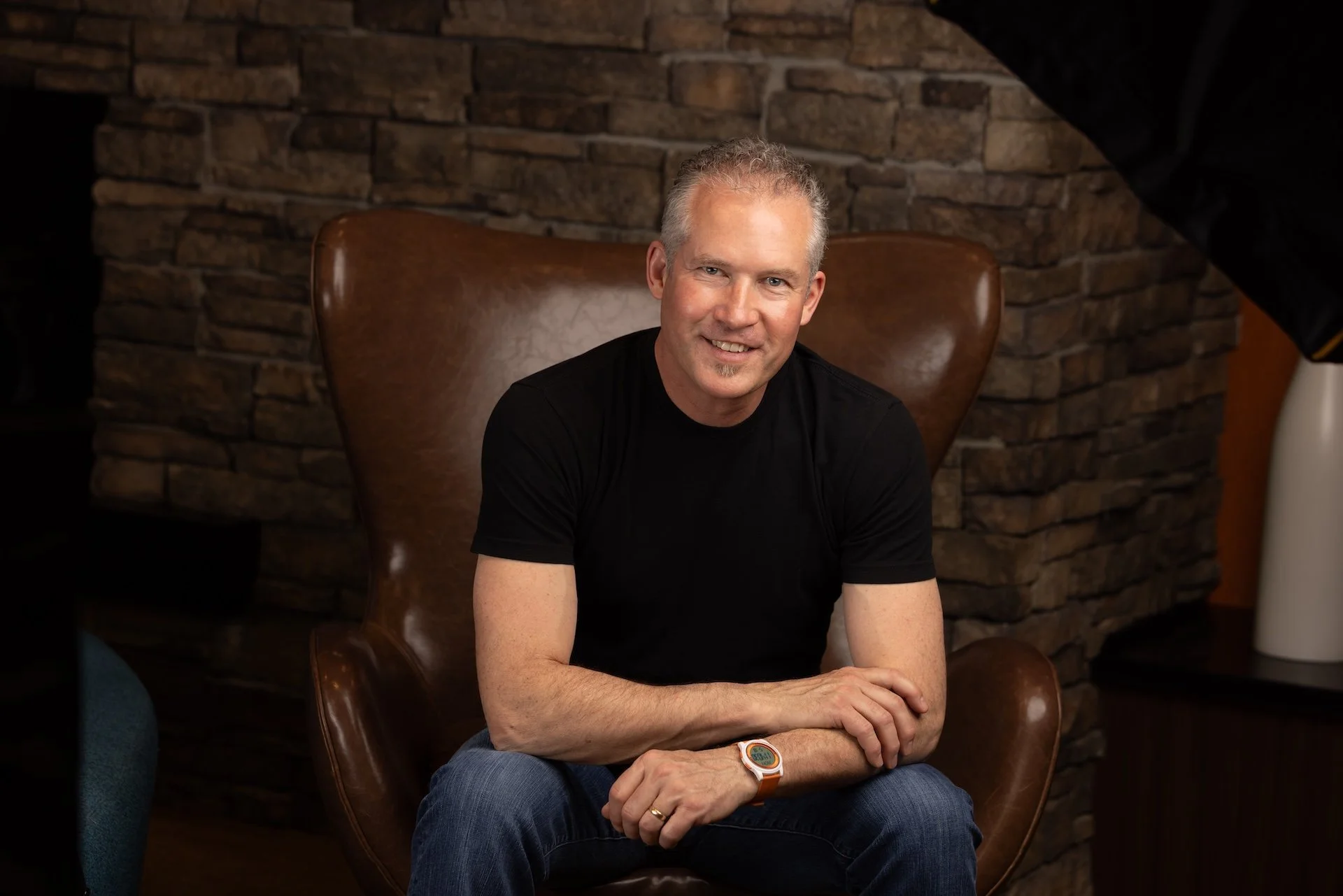 A smiling man with short gray hair and a goatee, wearing a black t-shirt, blue jeans, and an orange watch, sitting in a brown leather armchair in front of a brick wall.