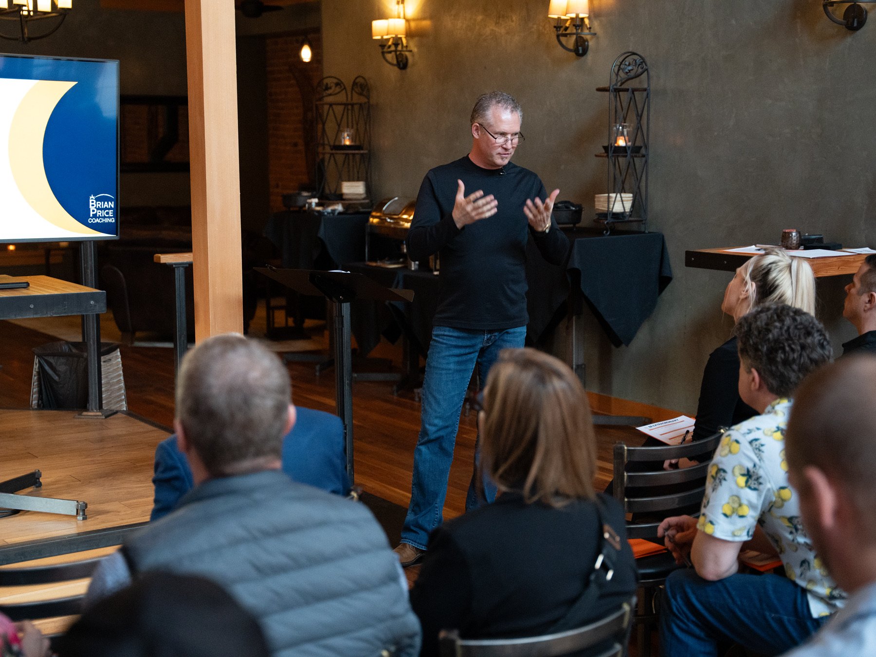 A man in black shirt and jeans giving a presentation to a group of people seated at tables in a cozy, dimly lit room with warm lamps, brick wall, and black metal decorative shelves. There is a screen on the left displaying a slide with the text 'BRIAN PRICE COACHING' and a large yellow crescent shape.