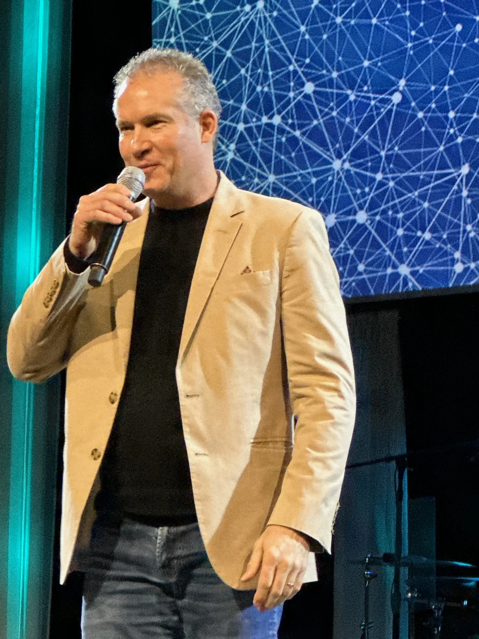 A man with gray hair and a tan blazer holding a microphone on stage with a blue abstract background.
