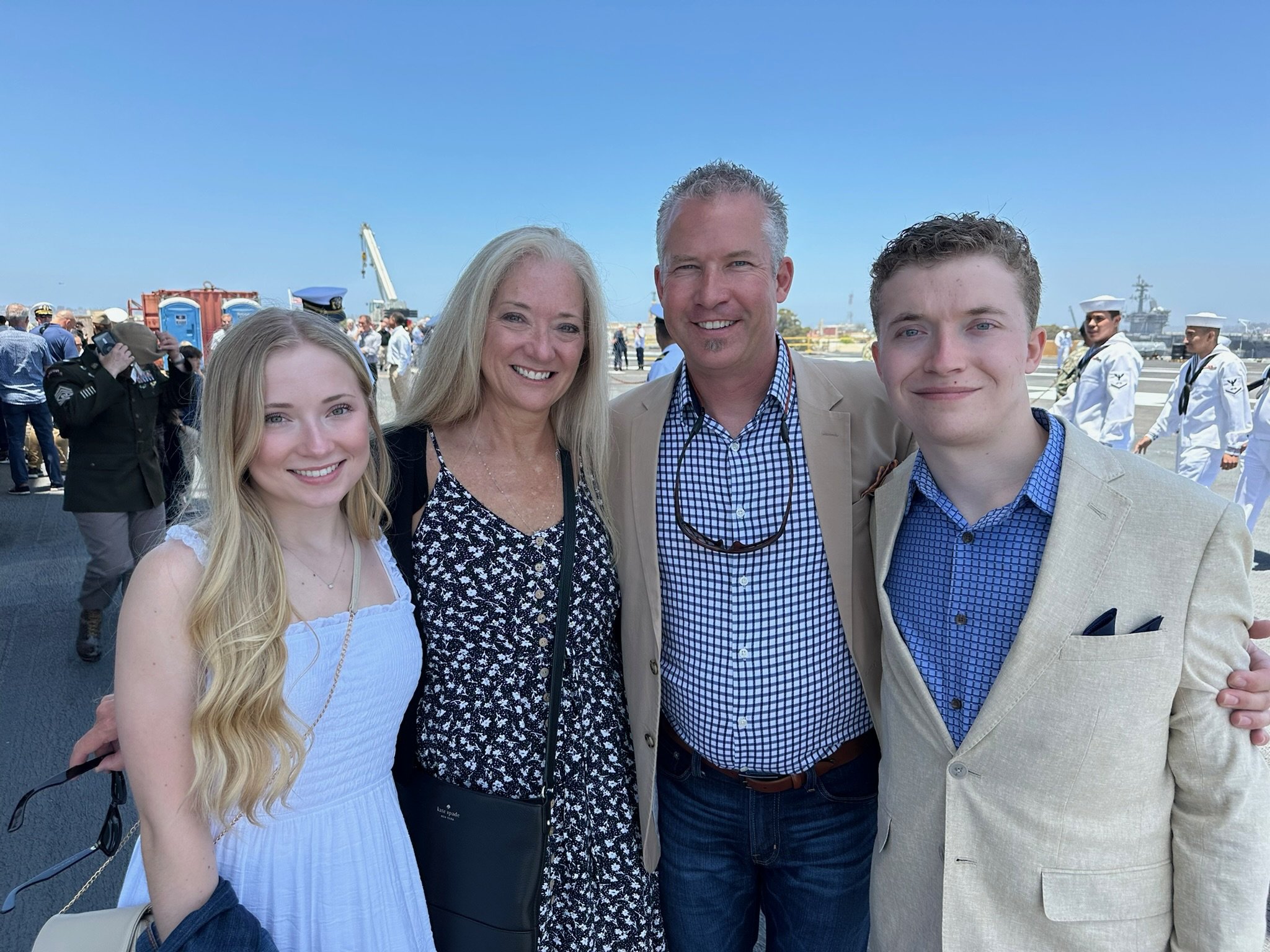 A group of four smiling people posing outdoors at a military event, with ships and military personnel in uniform visible in the background under a clear blue sky.