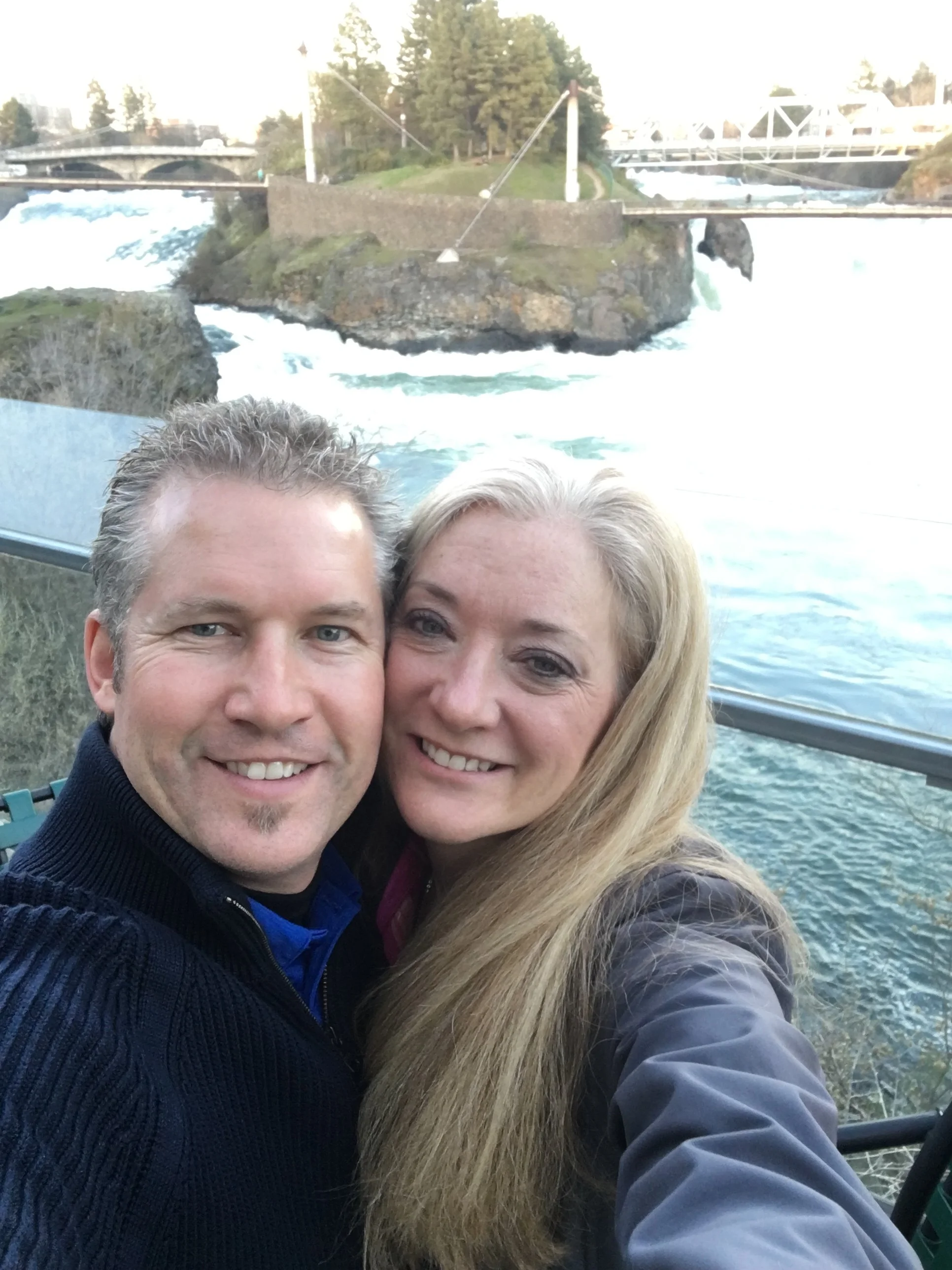 A smiling man and woman taking a selfie outdoors with a river, rocks, and bridges in the background.