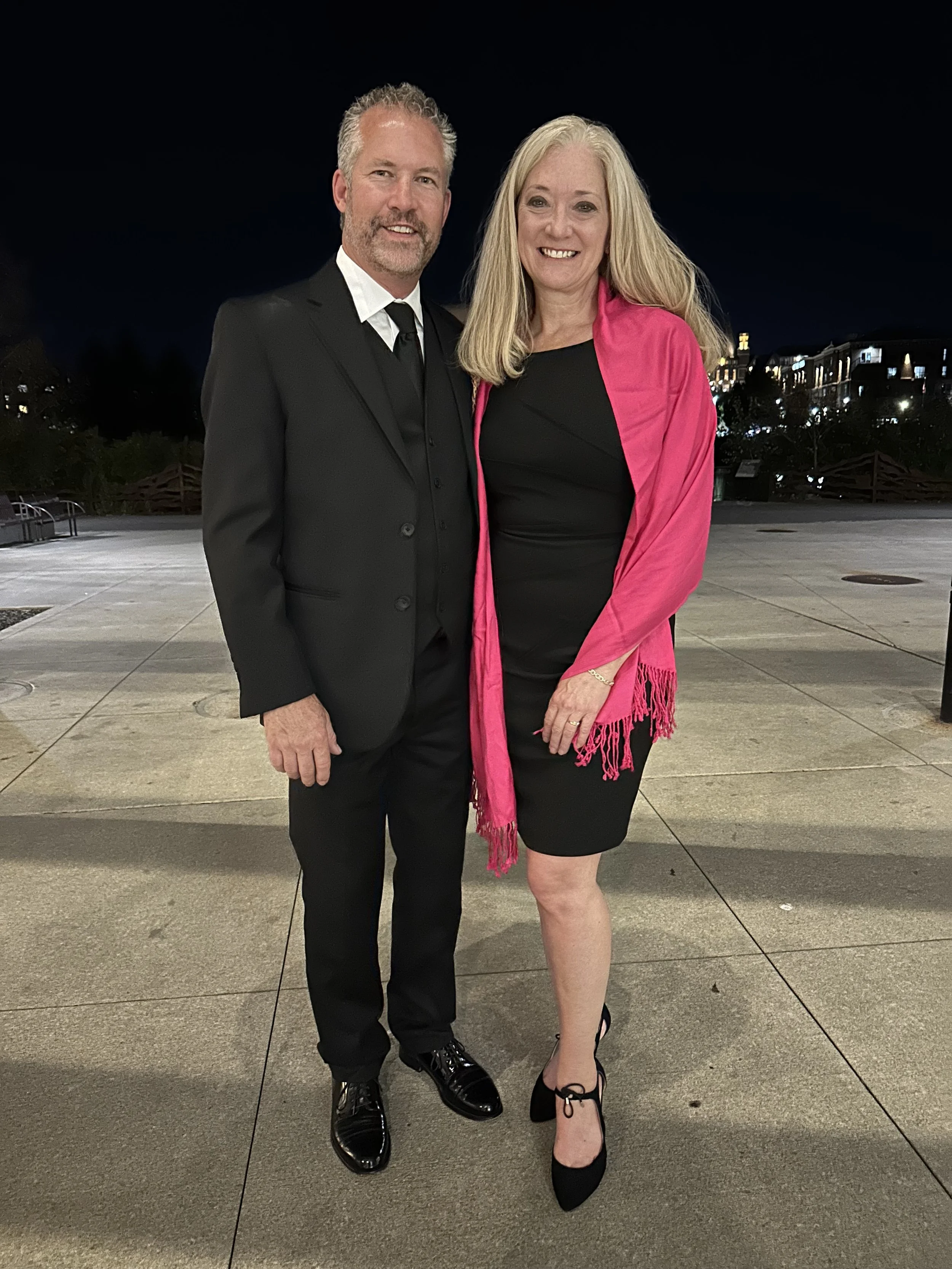 A man and a woman standing outdoors at night, dressed formally, smiling at the camera. The man is wearing a black suit with a tie and black dress shoes. The woman is wearing a black dress, pink shawl, and black high heels. There are city lights and buildings in the background.