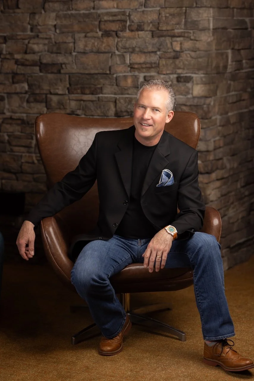 A middle-aged man with short gray hair sitting in a brown leather chair in front of a stone brick wall, wearing a black blazer, black shirt, blue jeans, and brown shoes, with a blue pocket square and a watch, smiling at the camera.