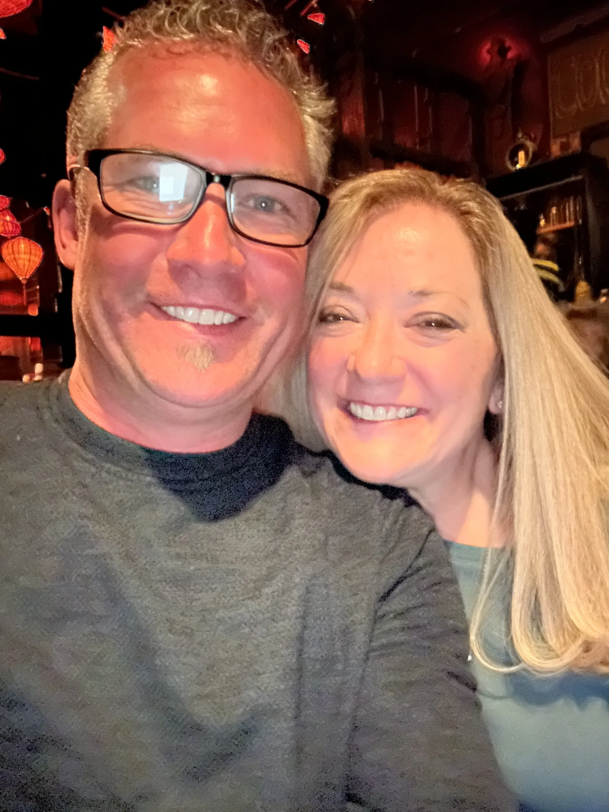 A close-up selfie of a man and a woman smiling, with the man on the left wearing glasses and the woman on the right with long blonde hair. The background appears to be a cozy indoor setting with warm lighting and decorative elements.