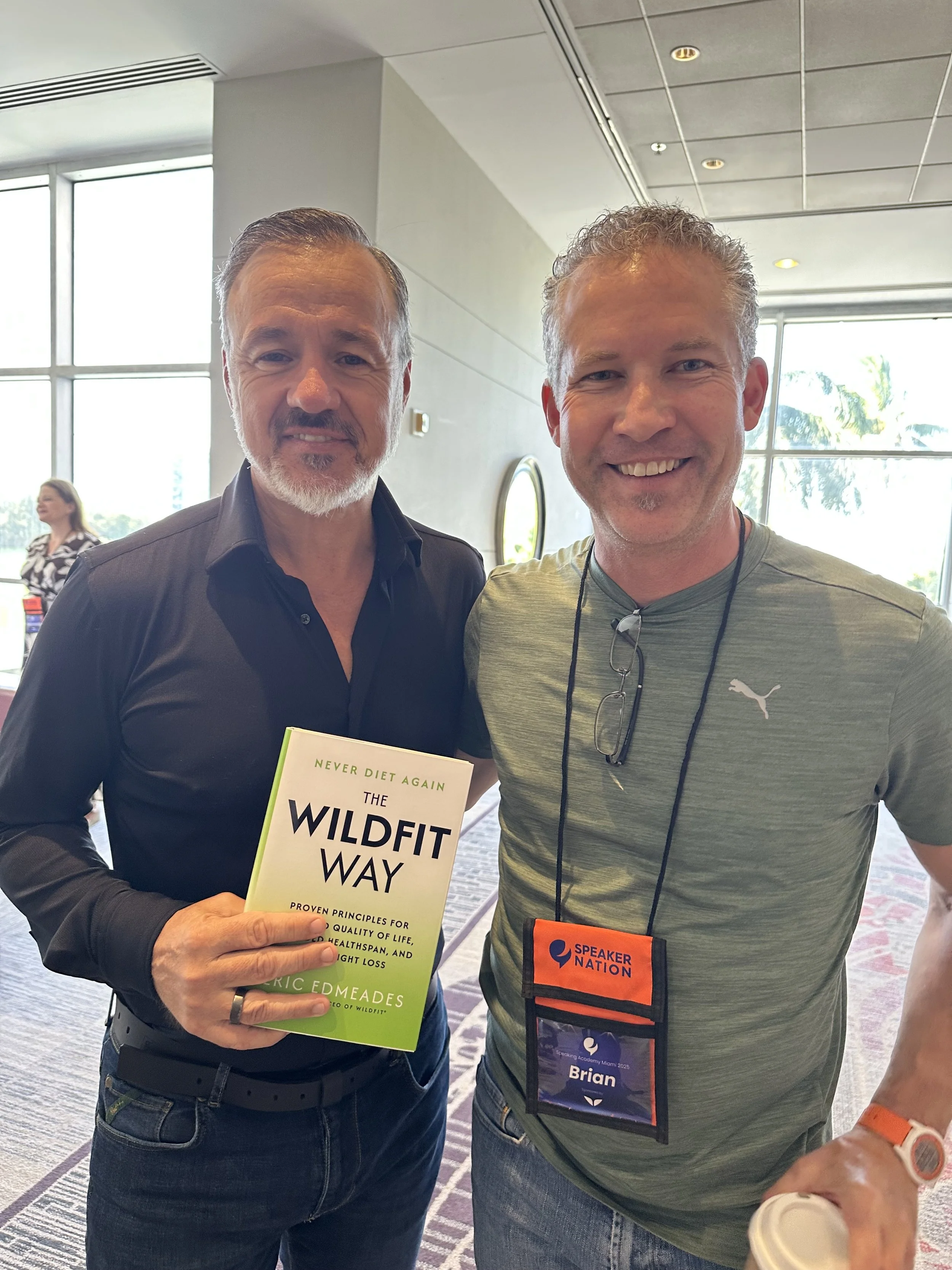 Two men standing together at an indoor event, one holding a book titled "The Wildfit Way" and wearing glasses around his neck, while the other is wearing a conference badge that reads "Brian". The background shows large windows and a woman in the distance.