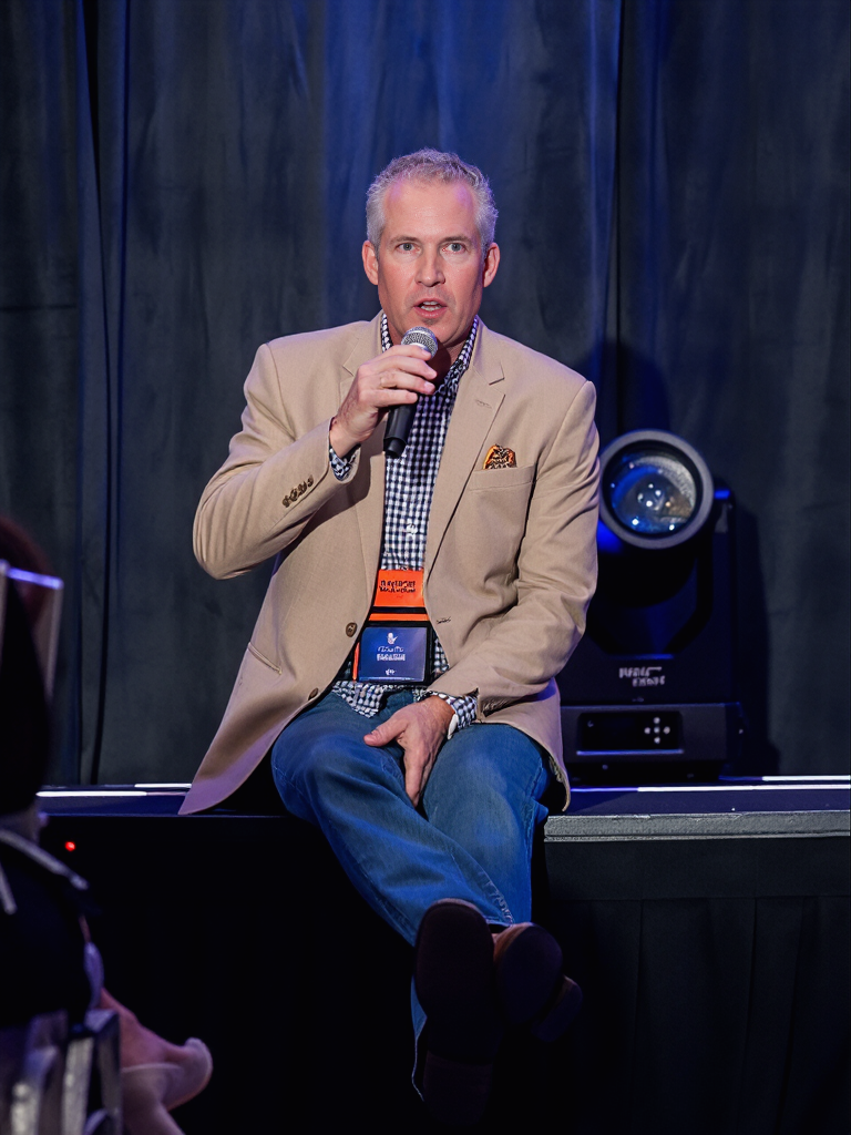 Man in beige blazer, checkered shirt, and jeans sitting on stage, holding a microphone and speaking at a conference.