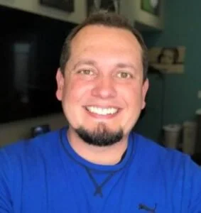 A smiling man with short hair and a goatee wearing a blue shirt, indoors.