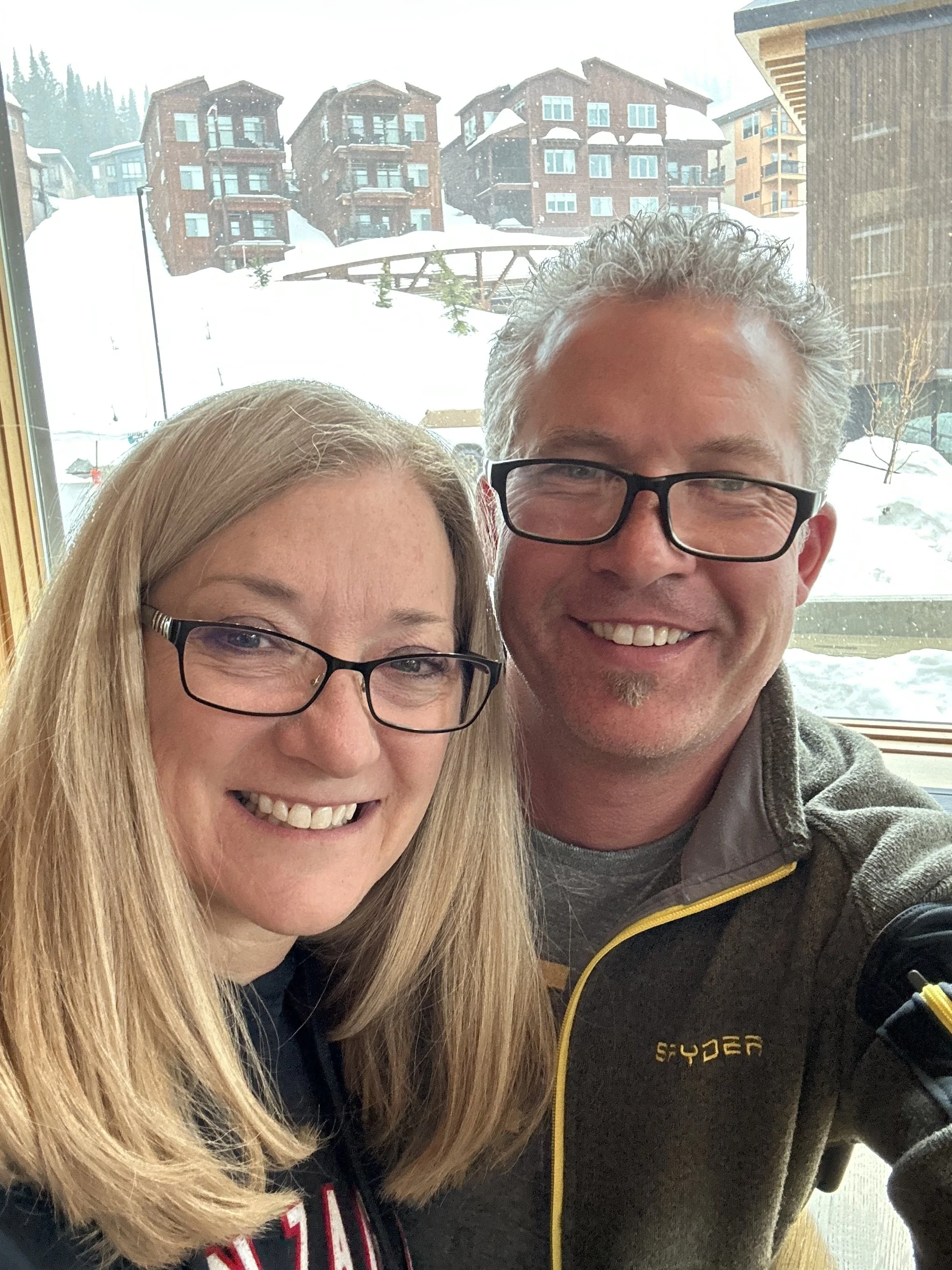 Close-up of a middle-aged couple smiling indoors with a snowy mountain village visible through the window behind them.