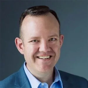 A smiling man with short dark hair wearing a blue blazer and light blue shirt against a gray background.