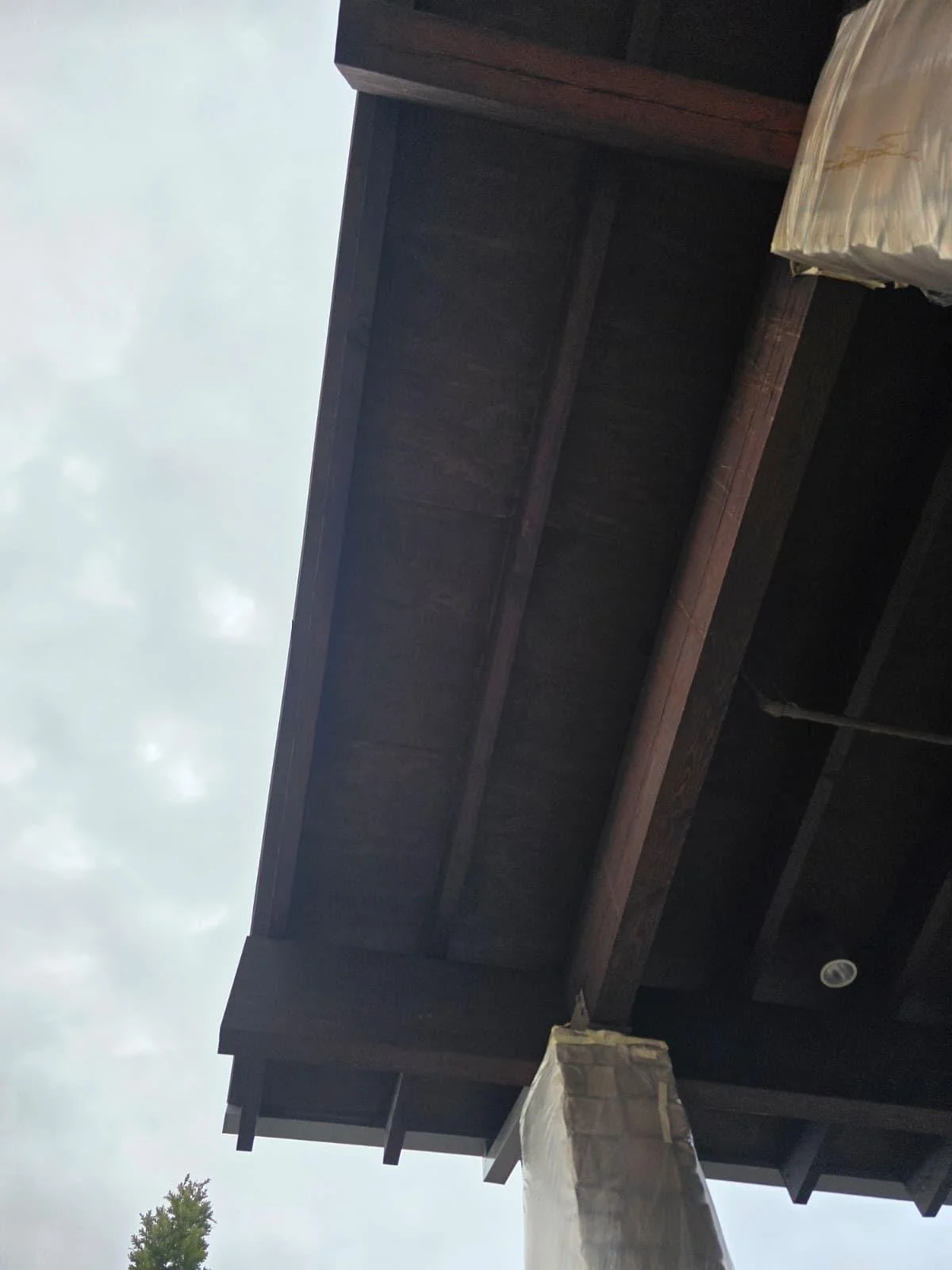 Close-up view of a dark wooden house exterior, showing the roof and a corner with a brick chimney against an overcast sky.