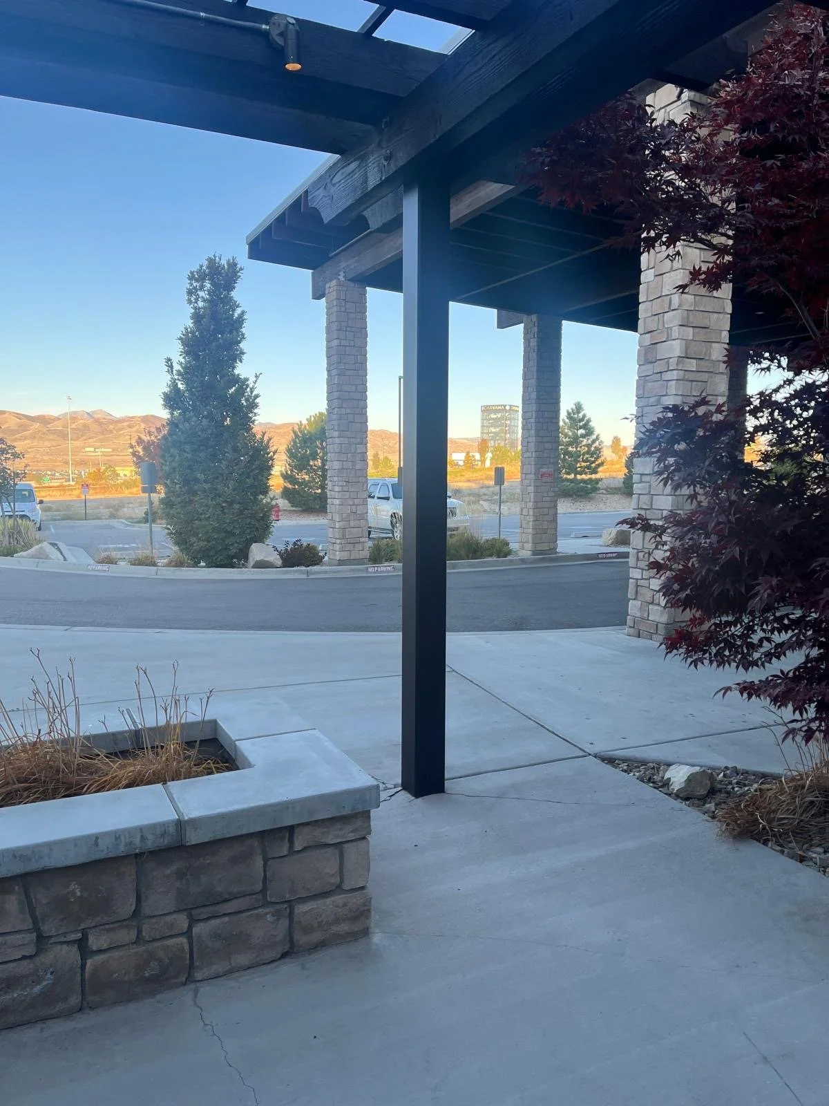 Outdoor scene with a concrete sidewalk, a stone planter with dried plants, a parking lot with cars, and a mountain range in the background, viewed from under a wooden and brick structure.