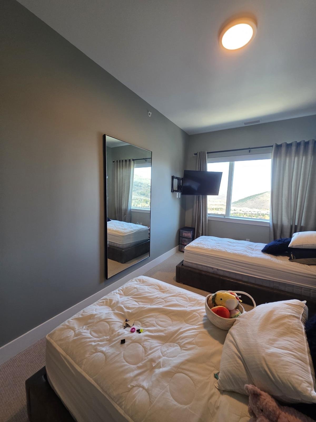 A bedroom with two unmade beds, a wall-mounted TV, a mirror, a window with curtains, and a small dresser. Items on one bed include a basket of plush toys and some small objects.