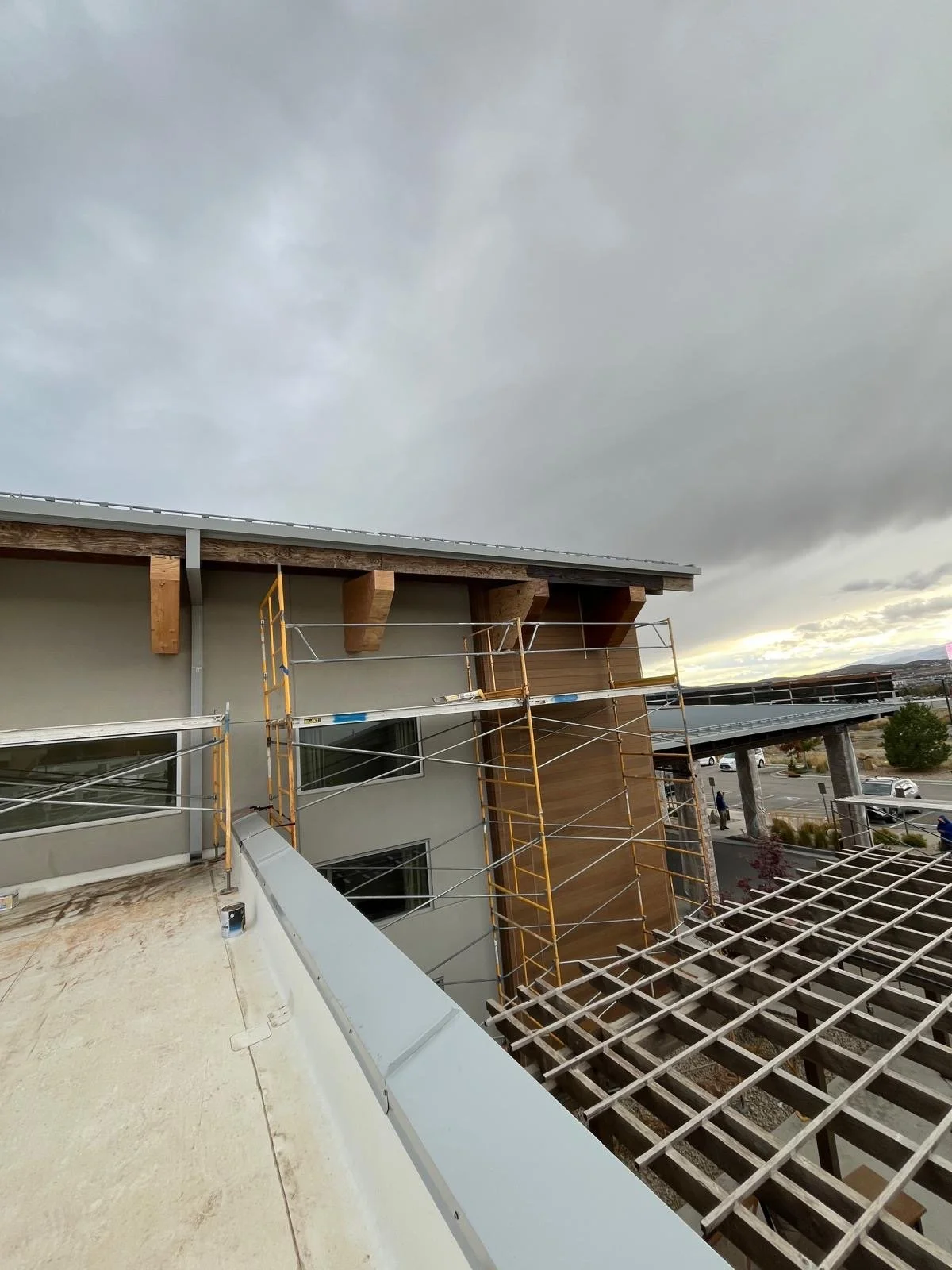 Construction workers installing roofing on a modern multi-story building with gray and wood facade, scaffolding, and a cloudy sky.