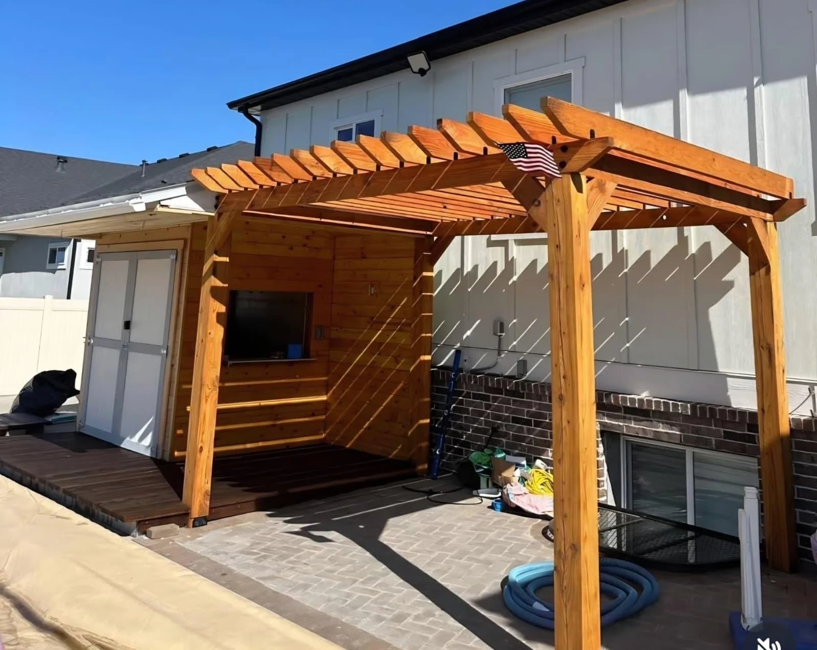 Backyard patio with a new wooden pergola and small wooden covered structure, with tools and hoses on the ground, and a blue sky overhead.