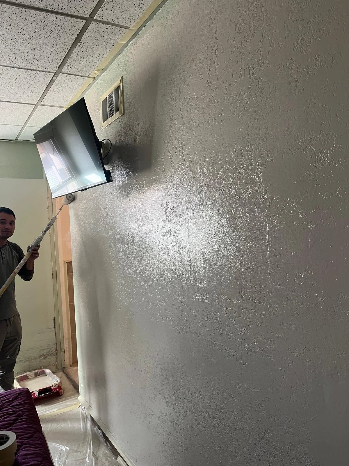 Person painting a wall with white paint, using a roller on an extension pole, in a room with a textured ceiling and a wall vent.