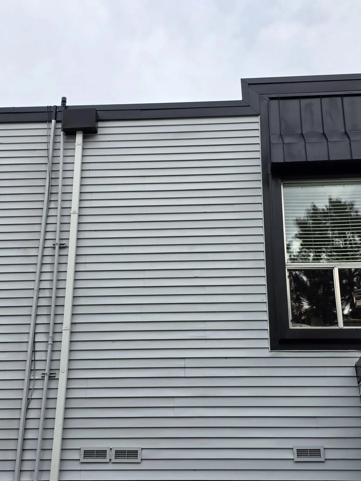 Close-up of a modern building's exterior wall with grey horizontal siding, black trim around a window, and visible electrical conduits.