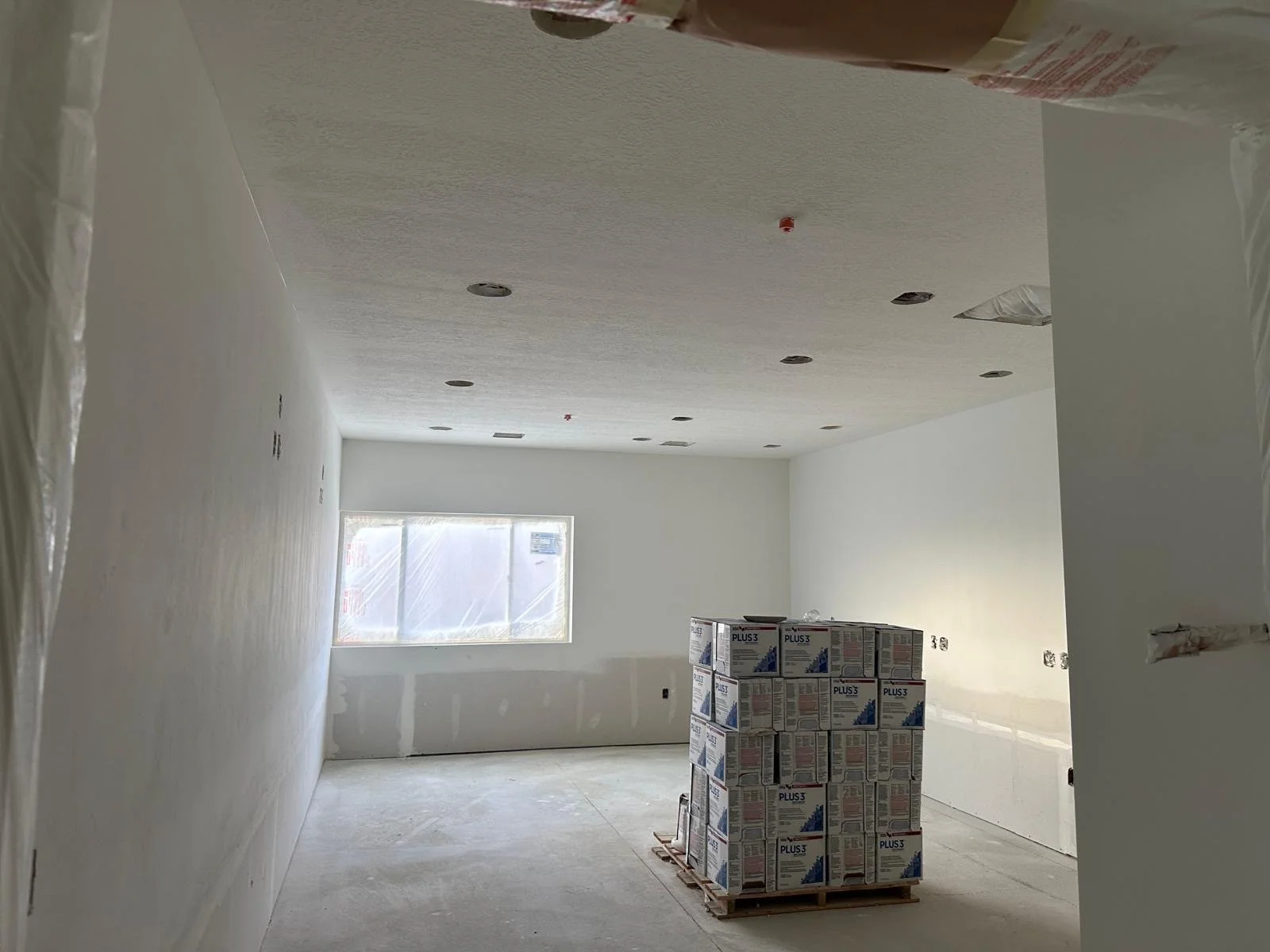Empty room under construction with white walls, a window covered with plastic, ceiling with recessed lighting, and a pallet of boxed materials in the center.
