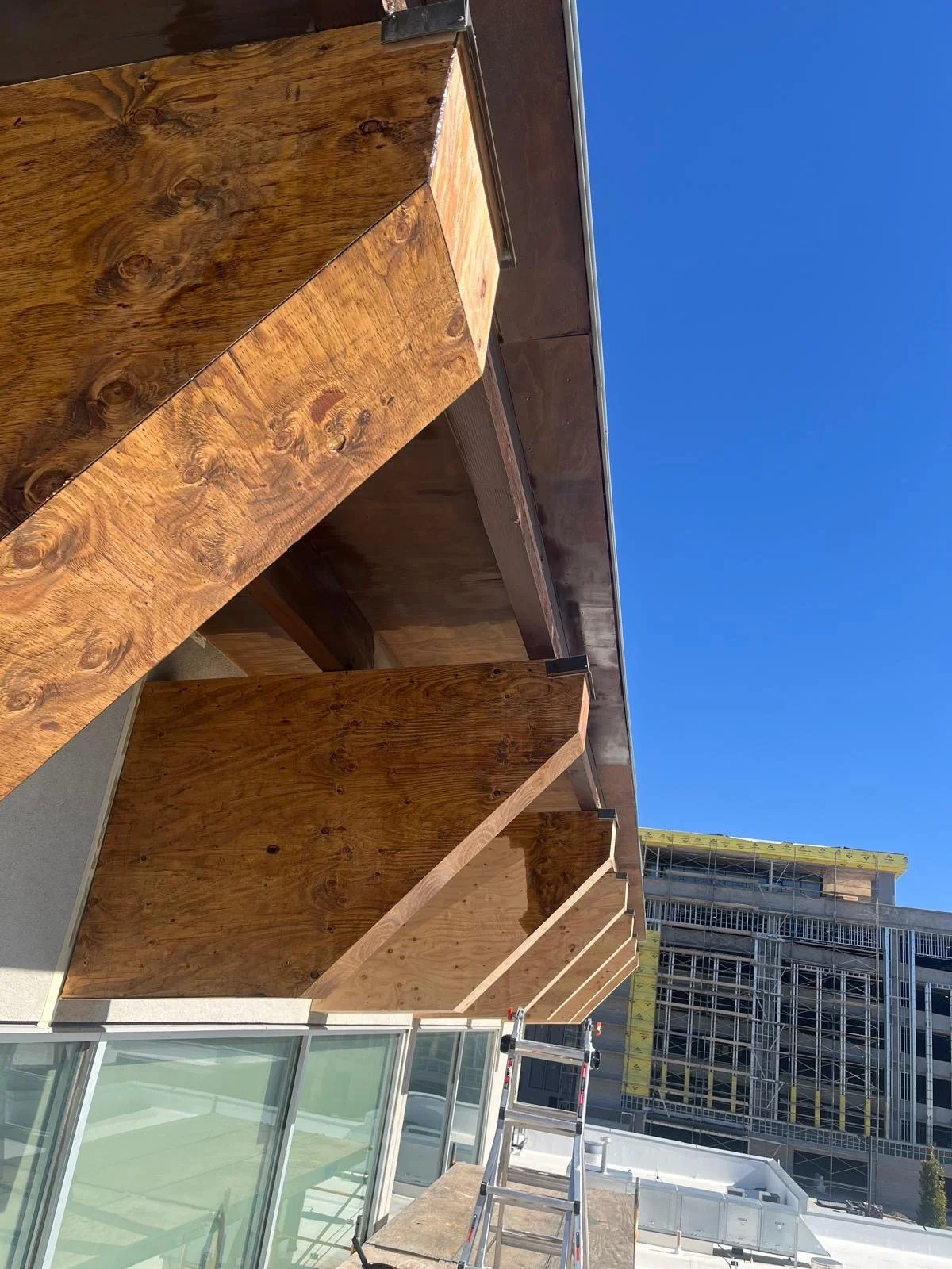 Construction site with a beginning of a building with large wooden panels and glass windows, scaffolding and a building under construction in the background, and a ladder in the foreground under a clear blue sky.