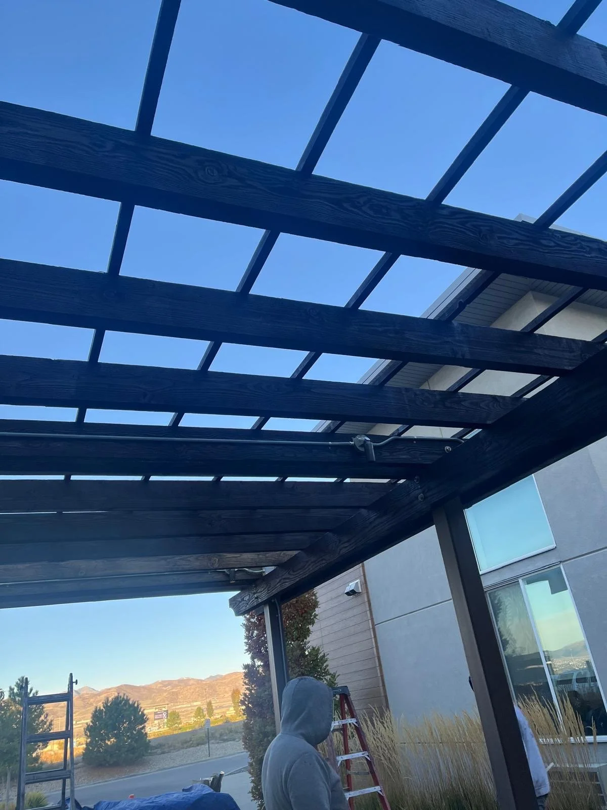 Person wearing a hoodie working on a wooden pergola outdoors with ladders and construction materials visible, desert landscape and mountains in the background.