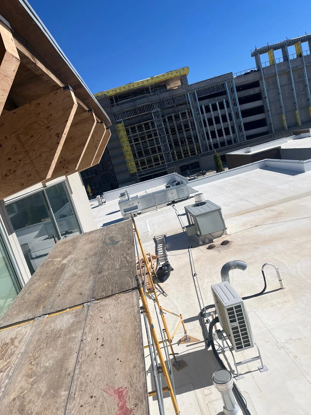 Construction site on a rooftop with scaffolding, ventilation units, and pipes, with an unfinished multi-story building in the background under a clear blue sky.