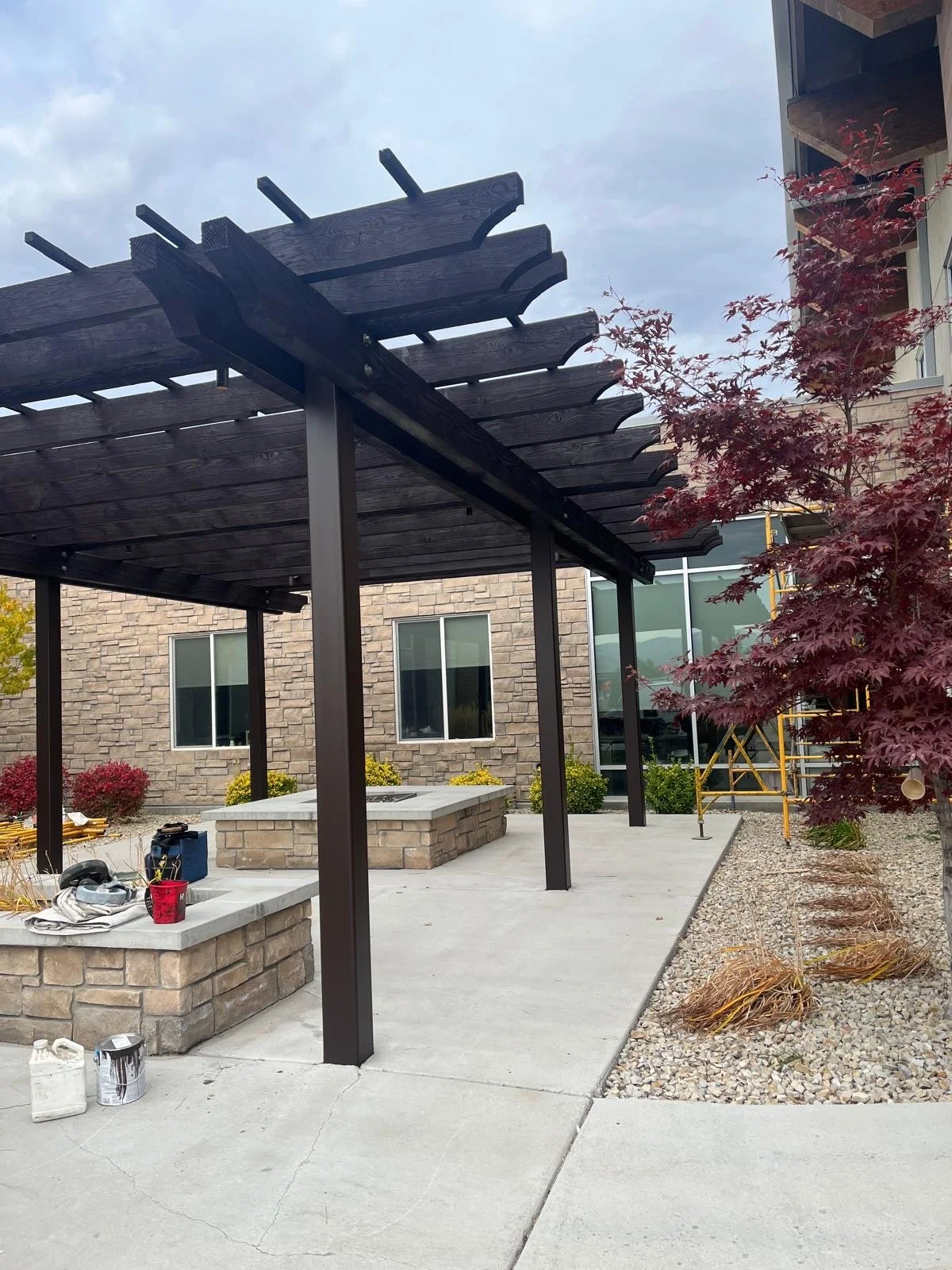 Construction site of a modern outdoor patio with a black wooden pergola, stone walls, concrete flooring, a small landscaped area with red and green foliage, and scaffolding nearby.