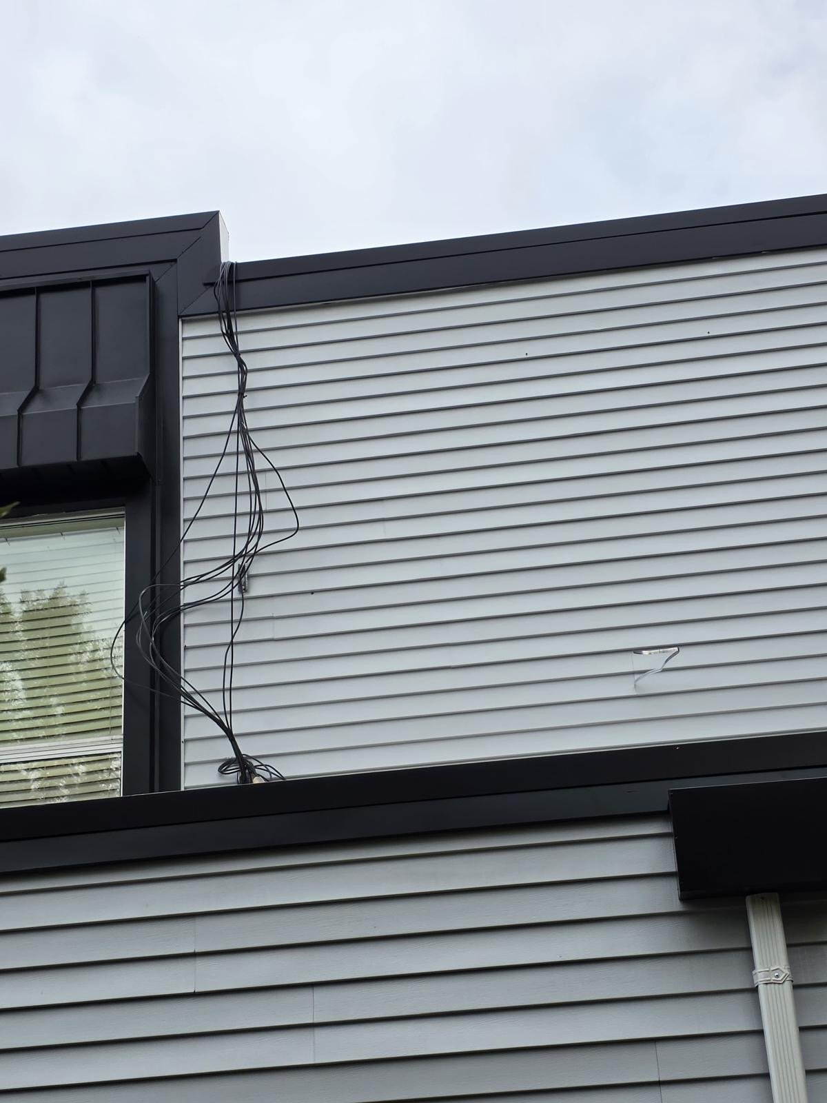 Close-up of the exterior of a modern building with black trim, light gray siding, and visible electrical wiring hanging from the top corner.