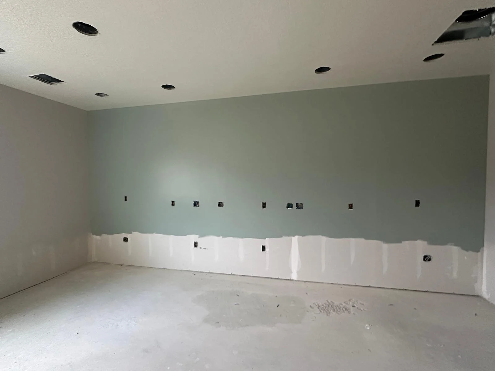 Empty room under construction with light green painted wall, multiple electrical outlets and holes in the ceiling for lights, unfinished flooring.