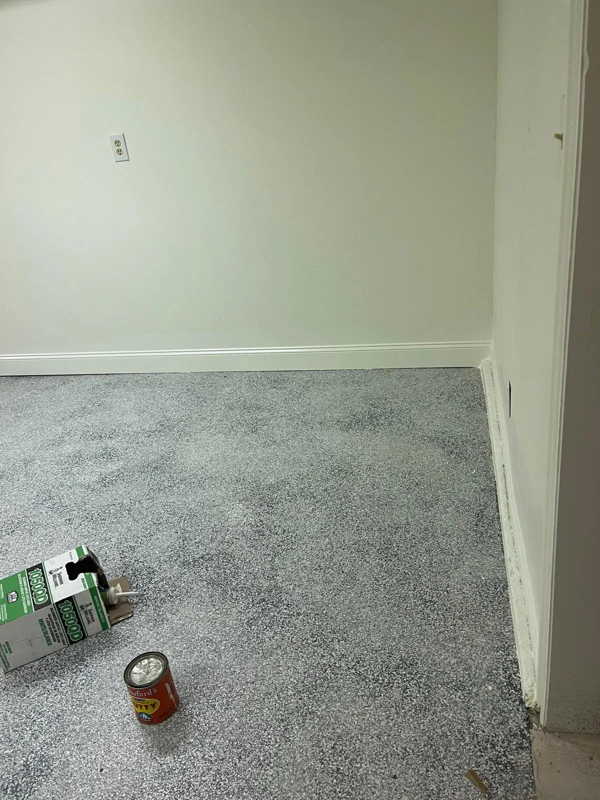 Empty room with light-colored wall, electrical outlet, white baseboard, and gray textured carpet. A can and a box of substance are on the floor.