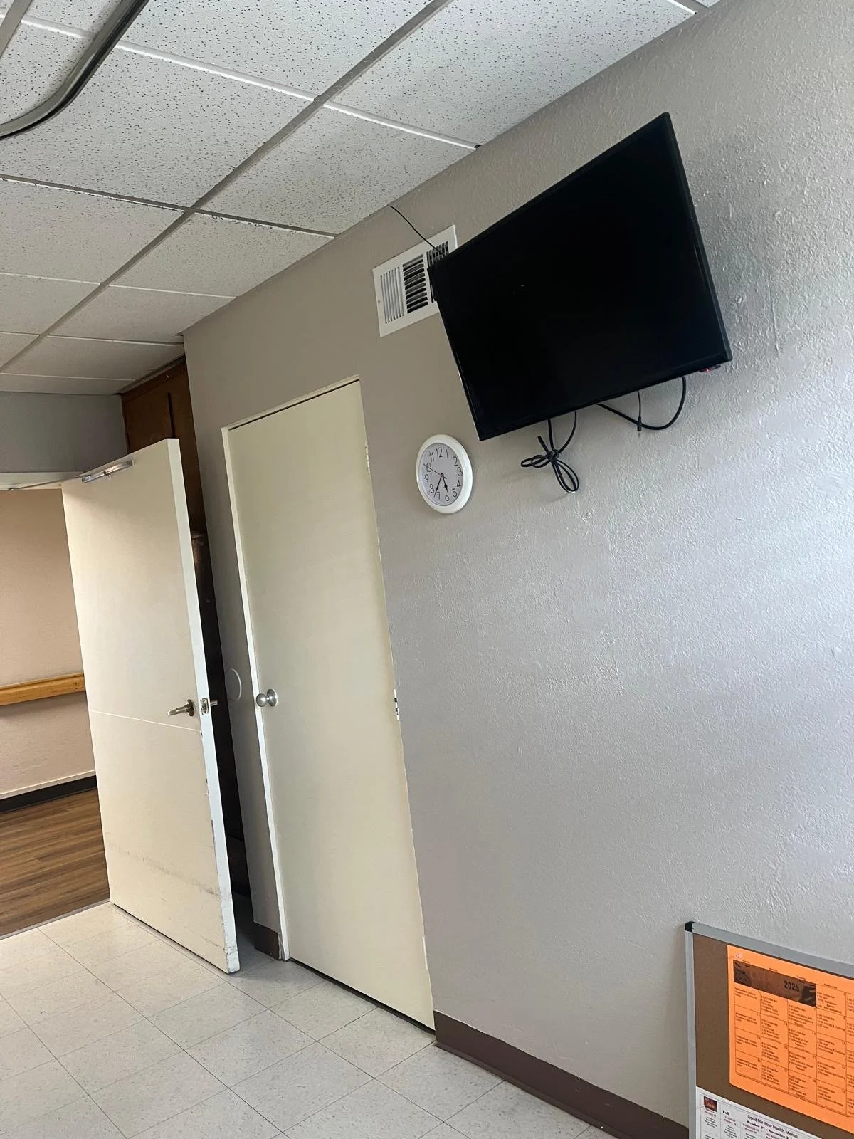 Hospital corridor with open door, closed door, wall clock showing 8:24, and mounted TV.