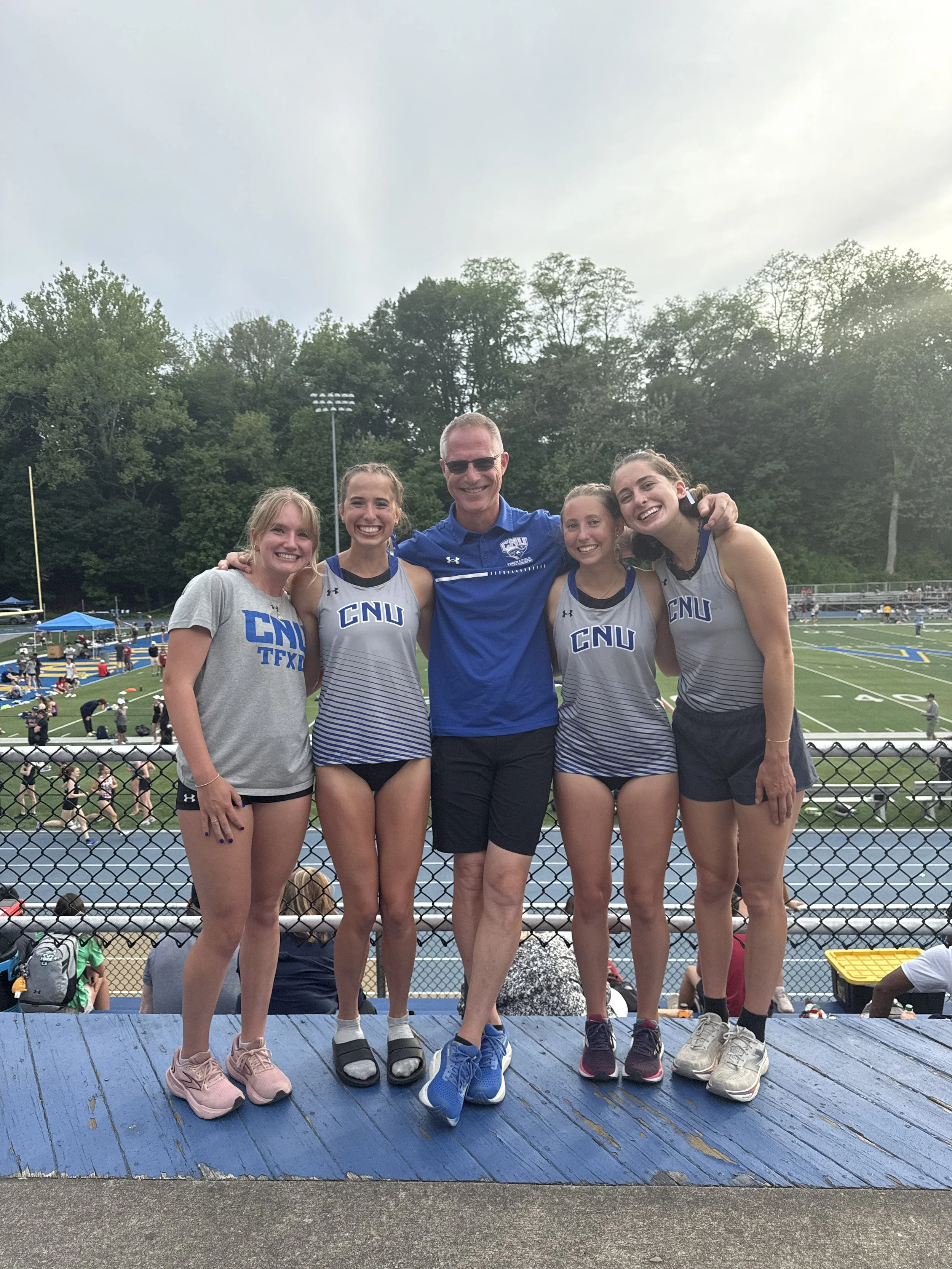 Group of four young women and a man, all smiling, standing on a blue platform on a sports field. The women are in athletic gear with 'CNU' written on their shirts, and the man is wearing sunglasses and a blue shirt. The background shows a football field with players warming up, trees, and overcast sky.