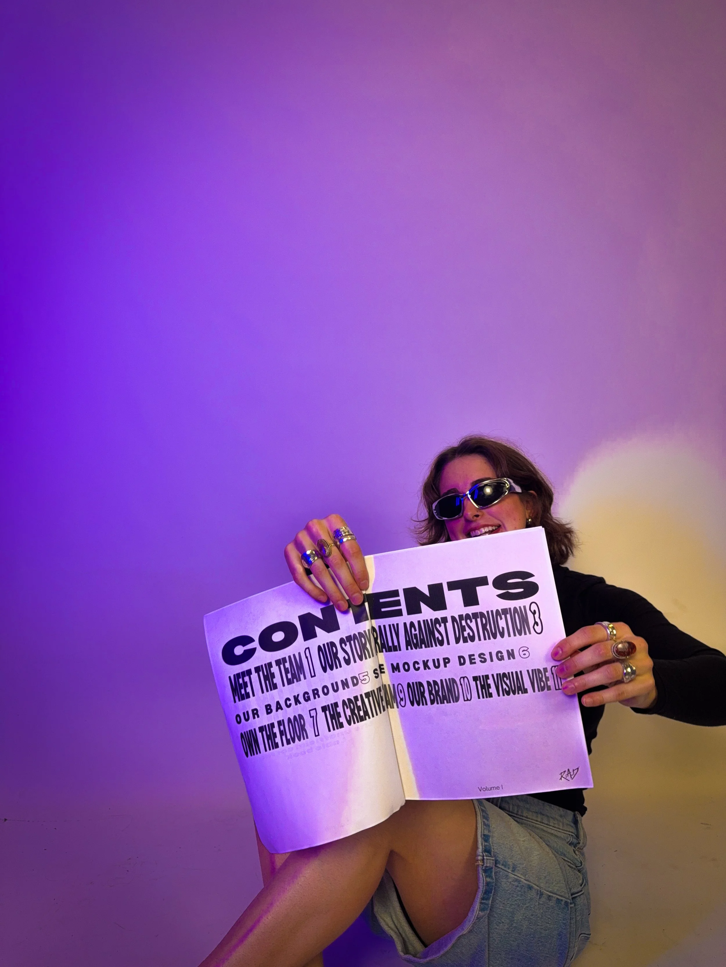 A woman wearing sunglasses and rings is sitting on the floor, smiling and holding an open magazine or booklet with bold text and various points listed, against a purple and yellow gradient background.