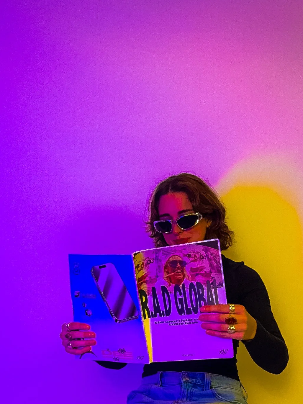 Woman with wavy hair, wearing sunglasses, reading a magazine titled 'R.A.D. GLOBAL' in front of a multicolored background.