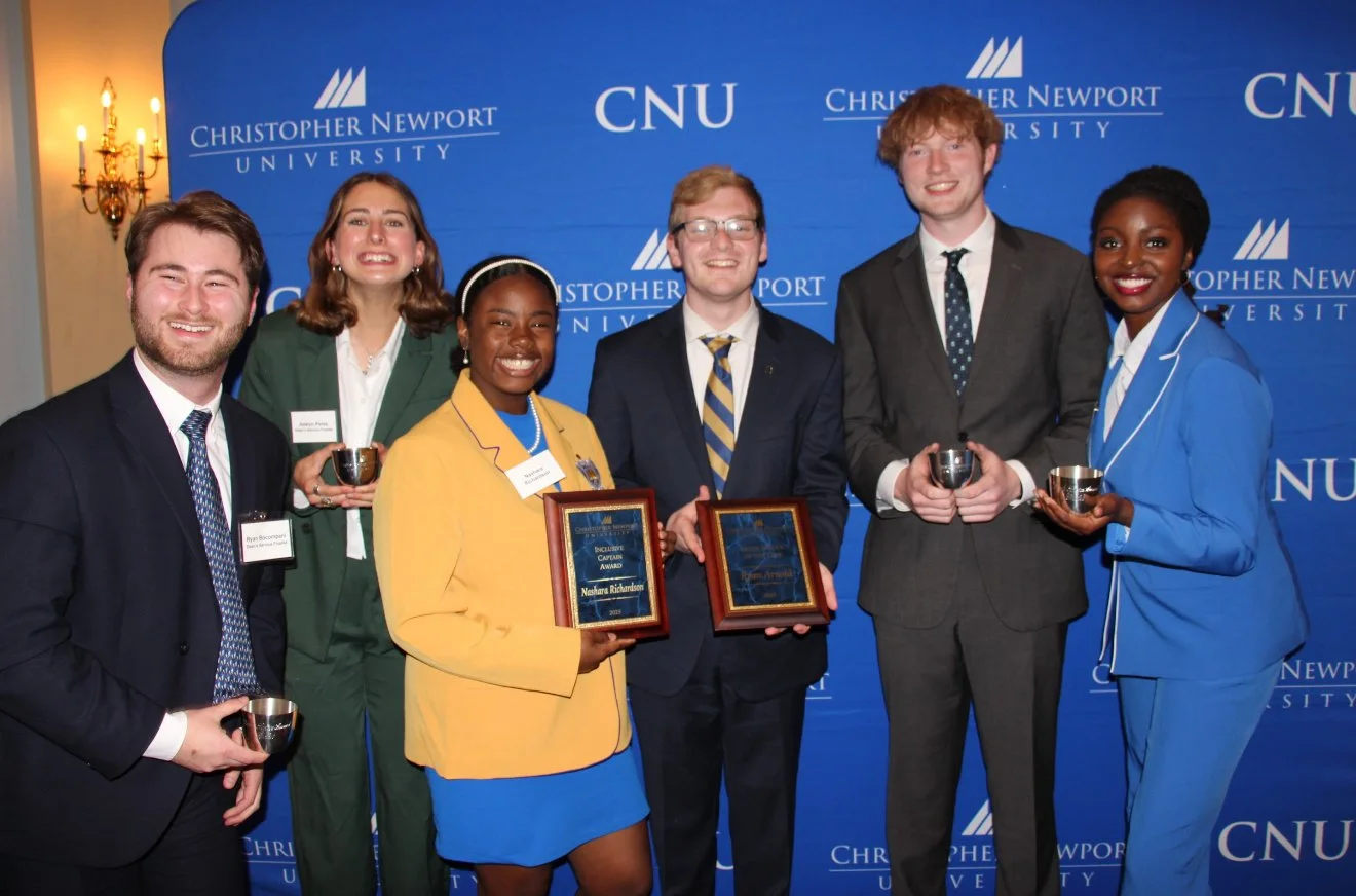 Group of seven diverse young adults standing in front of a blue backdrop with the Christopher Newport University logo, holding awards and smiling at the camera.
