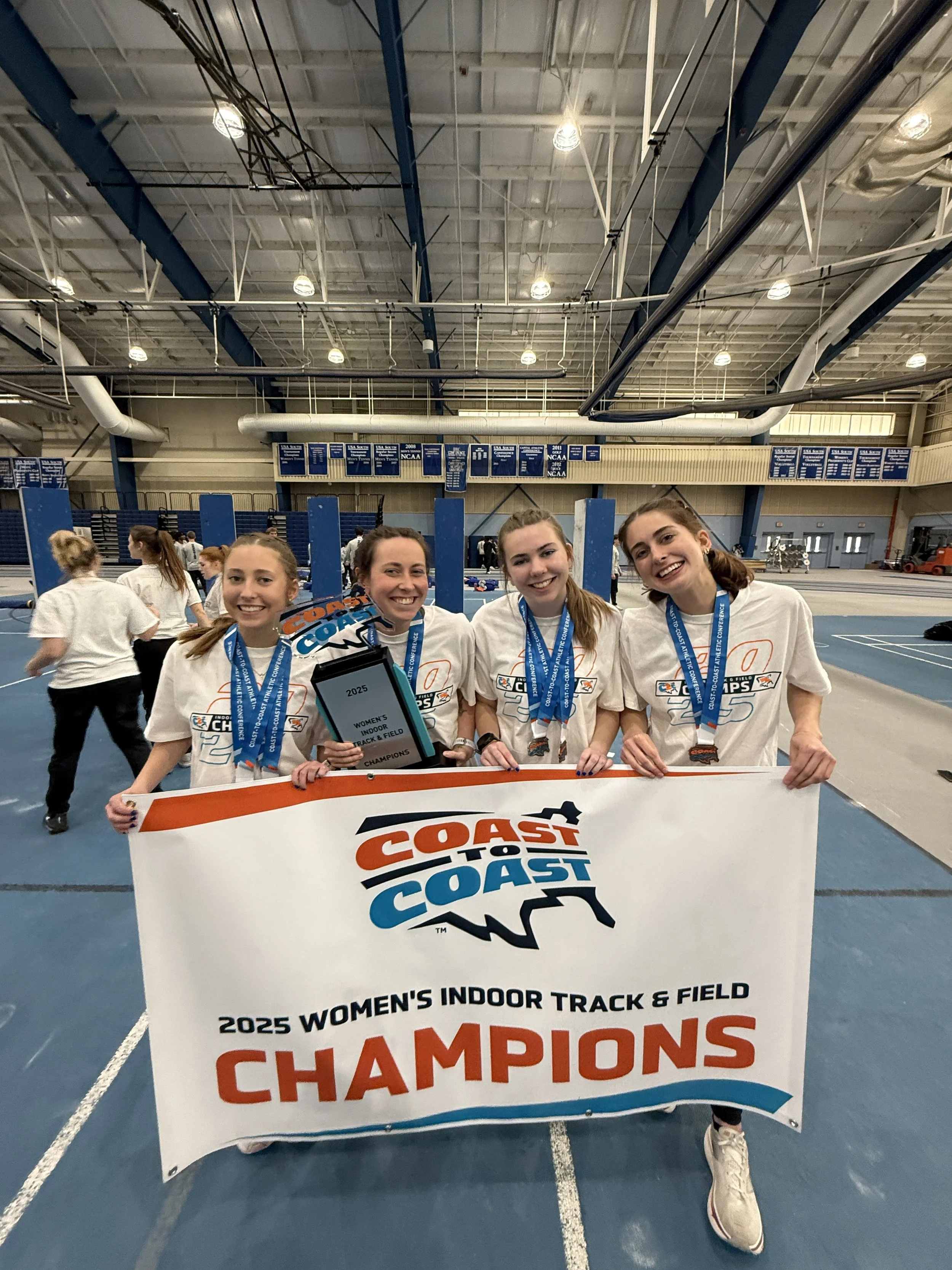 Four young women holding a championship banner at an indoor track and field facility, smiling, wearing medals, and celebrating their victory in the 2025 women's indoor track and field championship.