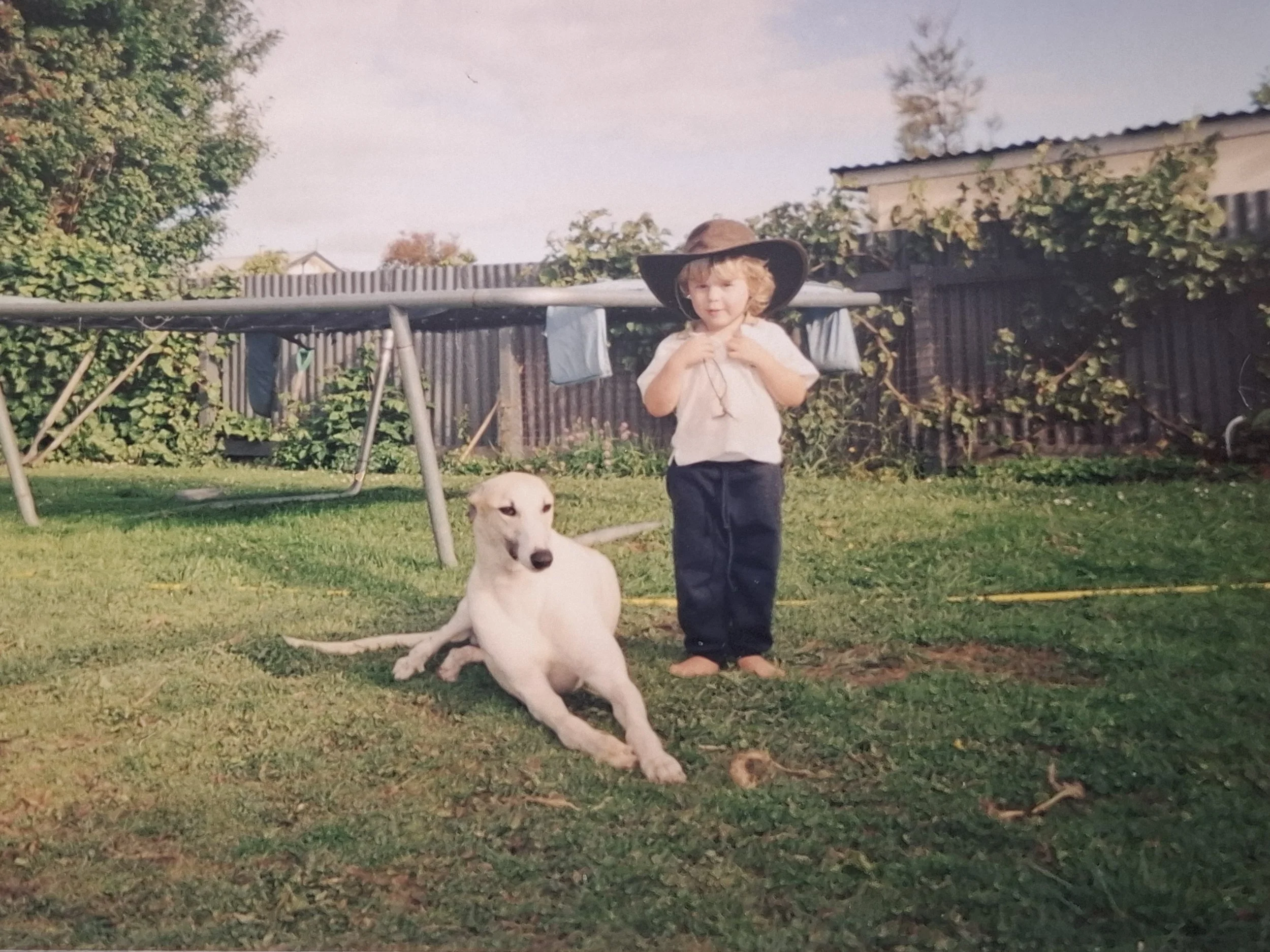 A young blonde boy wearing a large brown hat and beige shirt standing on grass in a backyard next to a white dog with black markings, with a wooden fence, garden, and trees in the background.