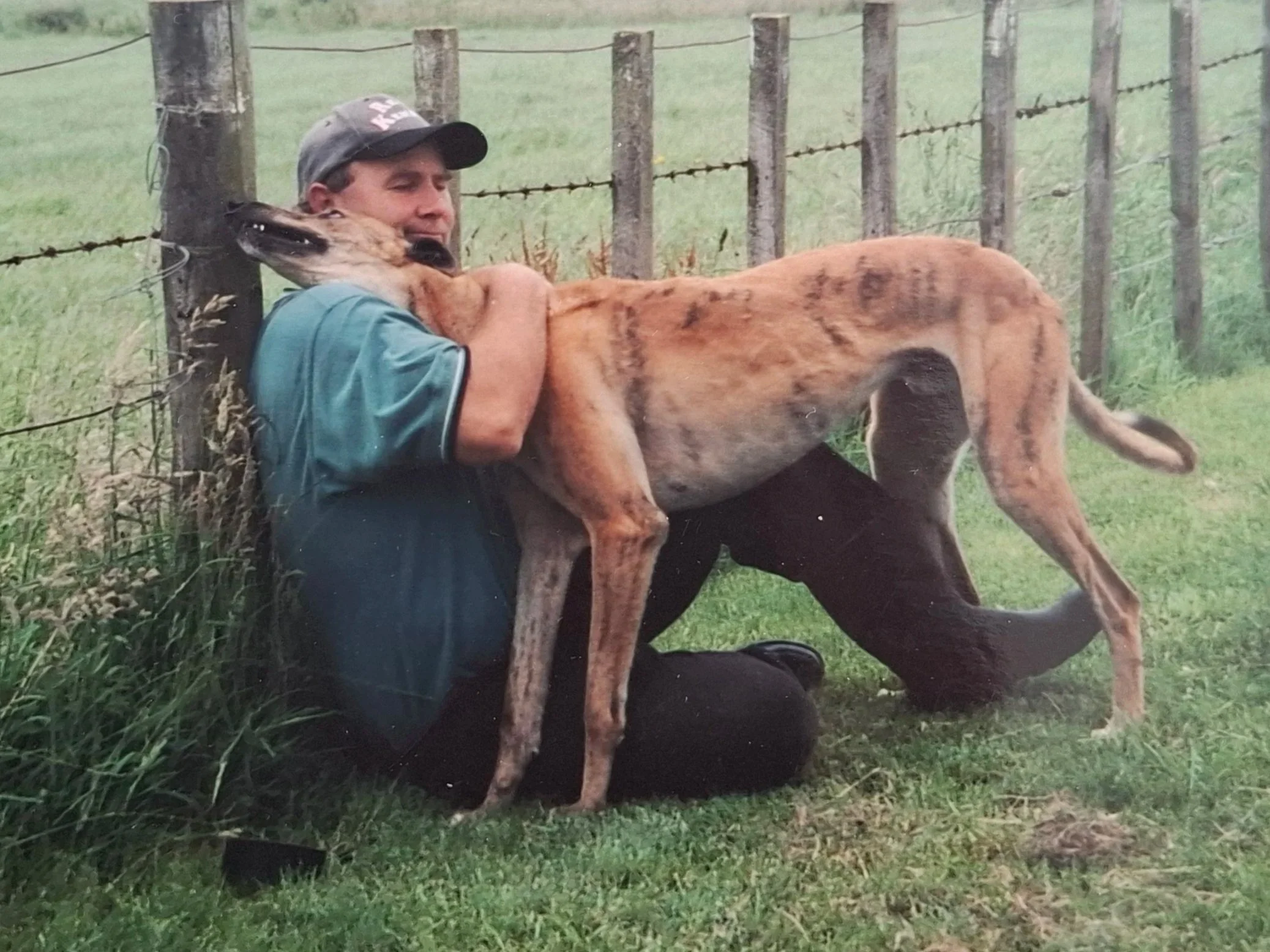 A man crouching on the grass leaning against a wooden fence post, hugging a large, tan and black striped dog. The dog appears to be resting its head on the man's shoulder. The background includes a field with a barbed wire fence and rolling hills.