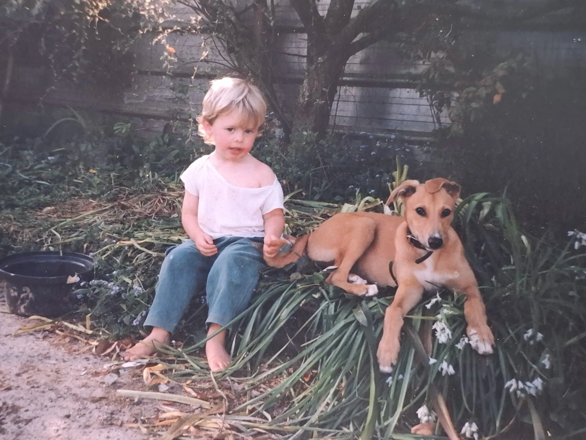 A young child with blond hair, wearing a loose white shirt and jeans, sits barefoot next to a tan and white dog on a patch of greenery with a fence in the background.