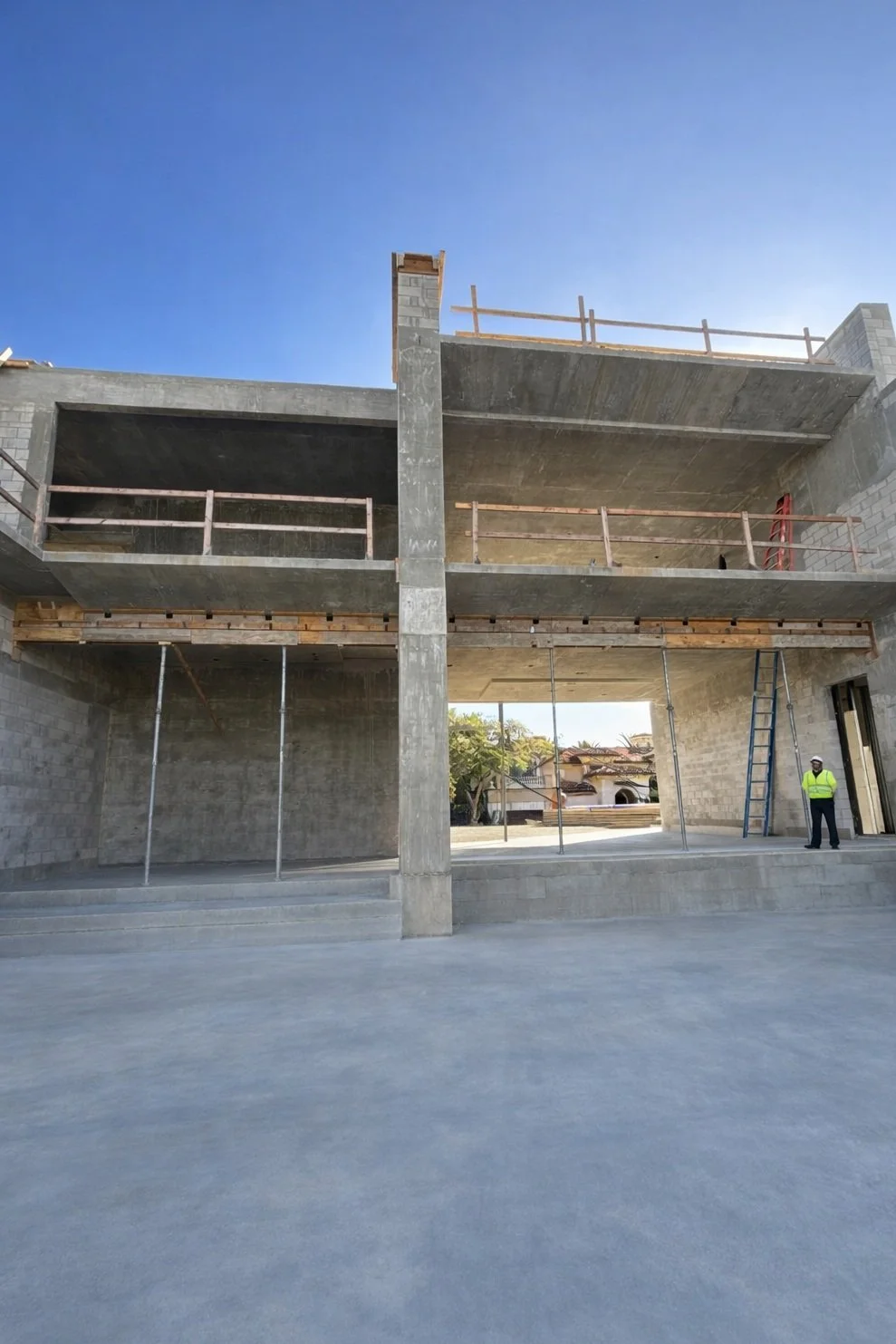 Construction site with a partially built multi-story concrete building under a clear blue sky, a worker wearing a safety vest and helmet standing near a ladder.