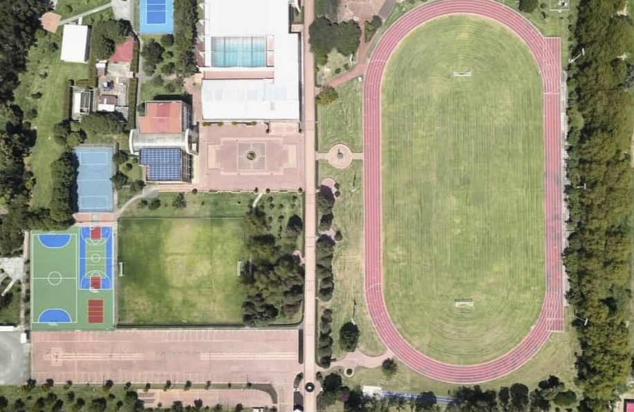 Aerial view of a sports complex with a large running track, soccer field, multiple tennis courts, a swimming pool, and various buildings surrounded by greenery.