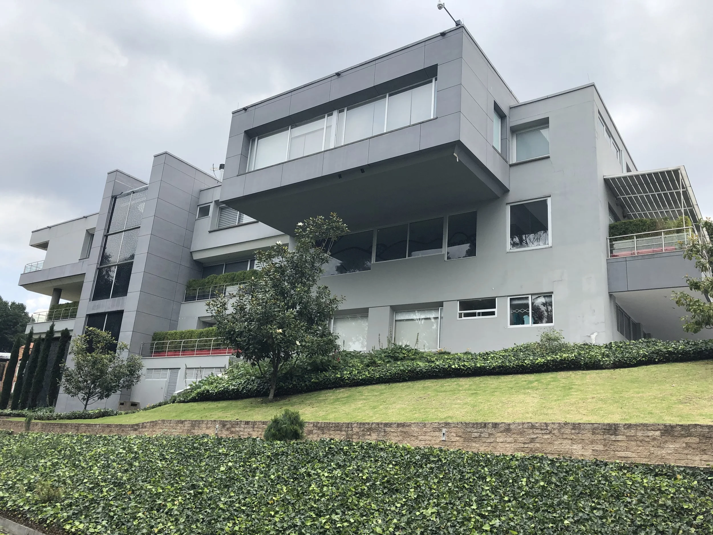 A modern multi-story residential building with large windows and balconies, surrounded by greenery and landscaped lawns, under a cloudy sky.